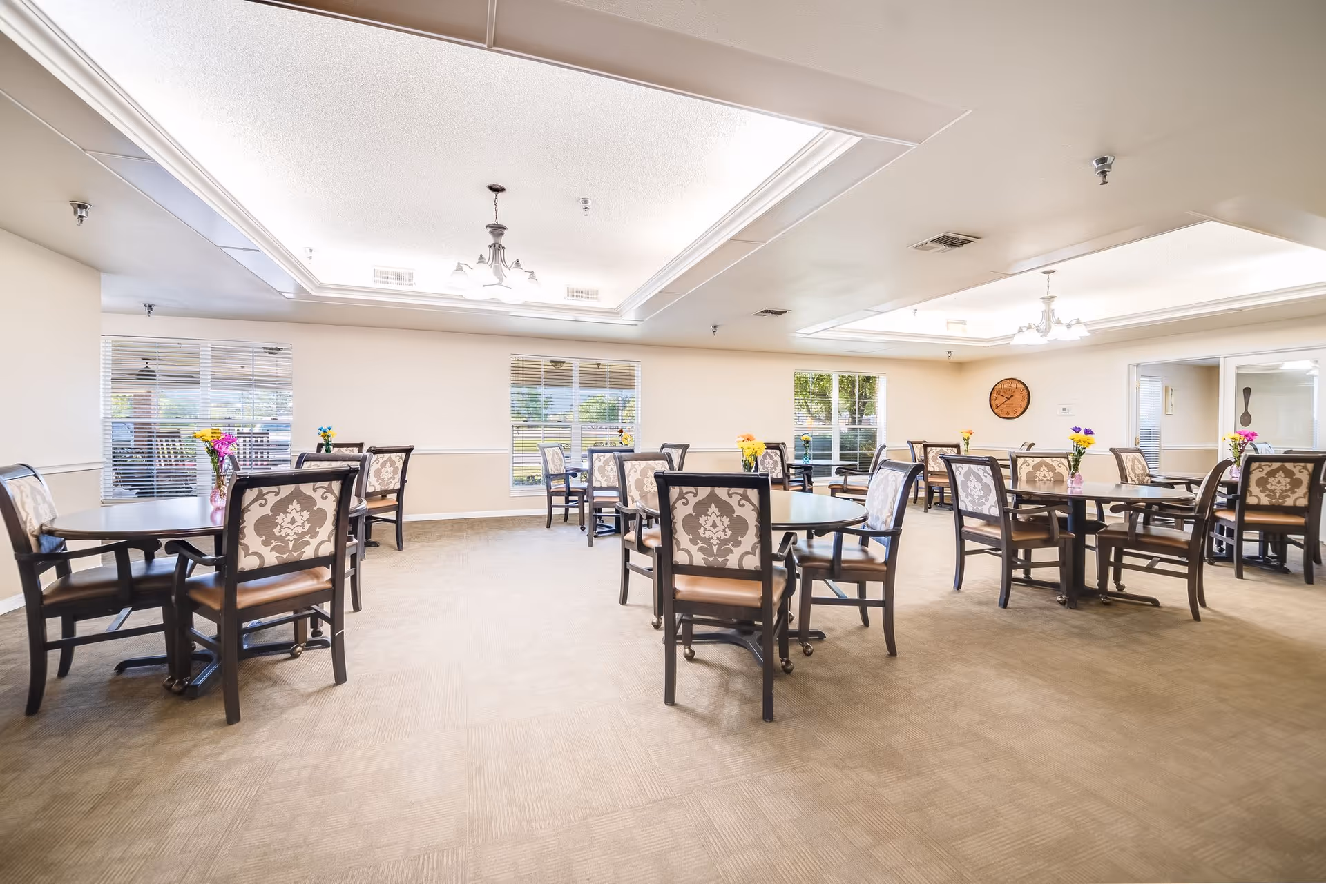 A bright and spacious dining room with multiple round tables and cushioned chairs arranged neatly. Each table has a small vase with colorful flowers. Large windows allow natural light to fill the room, and there are ceiling lights and a wall clock visible.