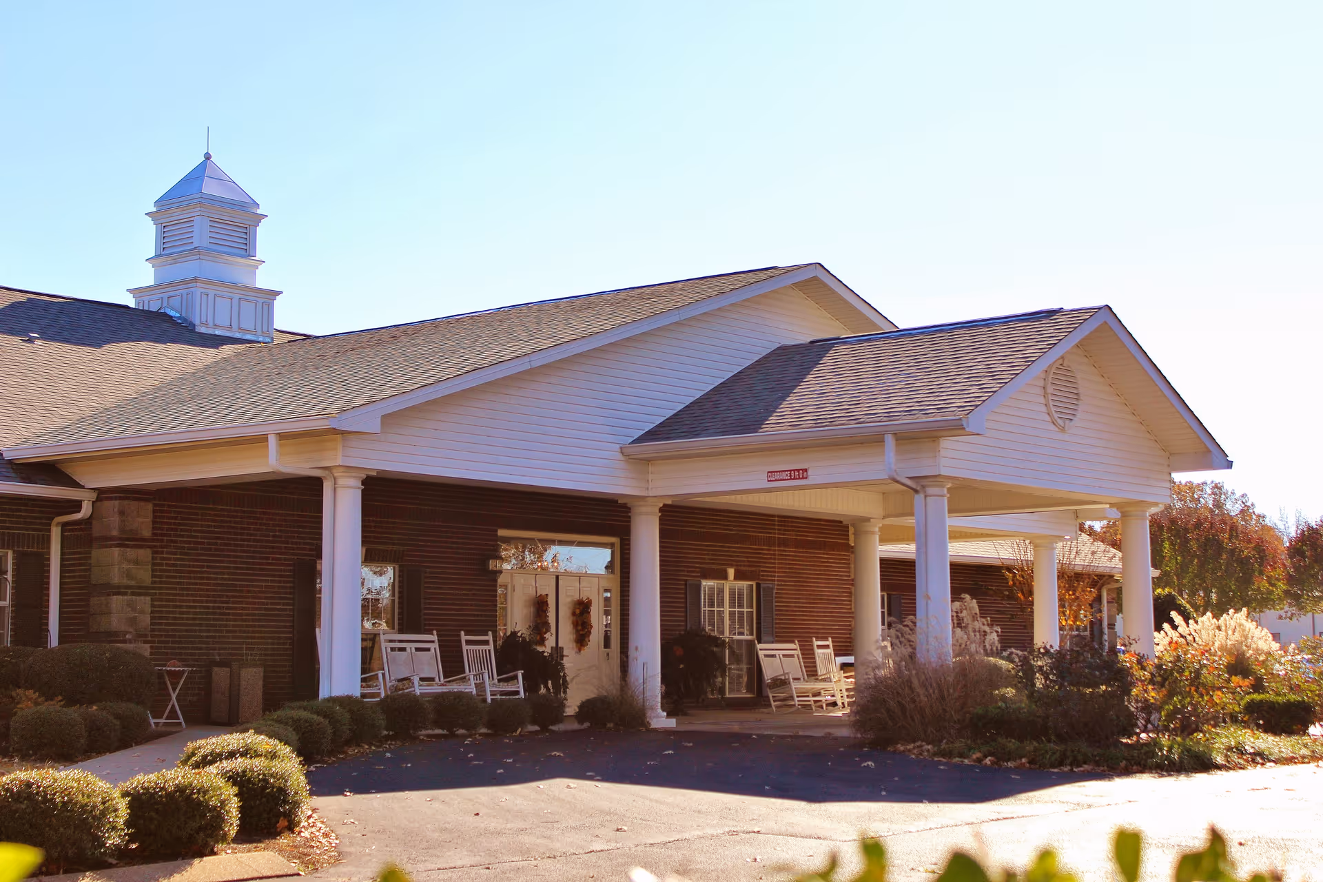 Exterior view of Sugar Creek Senior Living facility showing a covered entrance with white columns, rocking chairs on the porch, brick walls, and landscaped bushes and plants under a clear sky.