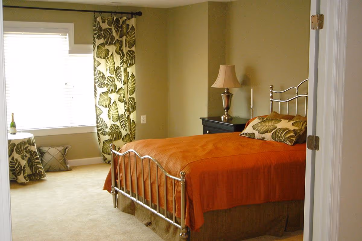 A bedroom with a metal frame bed covered with an orange bedspread and a pillow with a green leaf pattern. There is a black nightstand with a lamp and a candle on it. A window with white blinds and curtains with a green leaf pattern is on the left side. A small round table covered with a matching green leaf patterned cloth and a cushion are near the window.