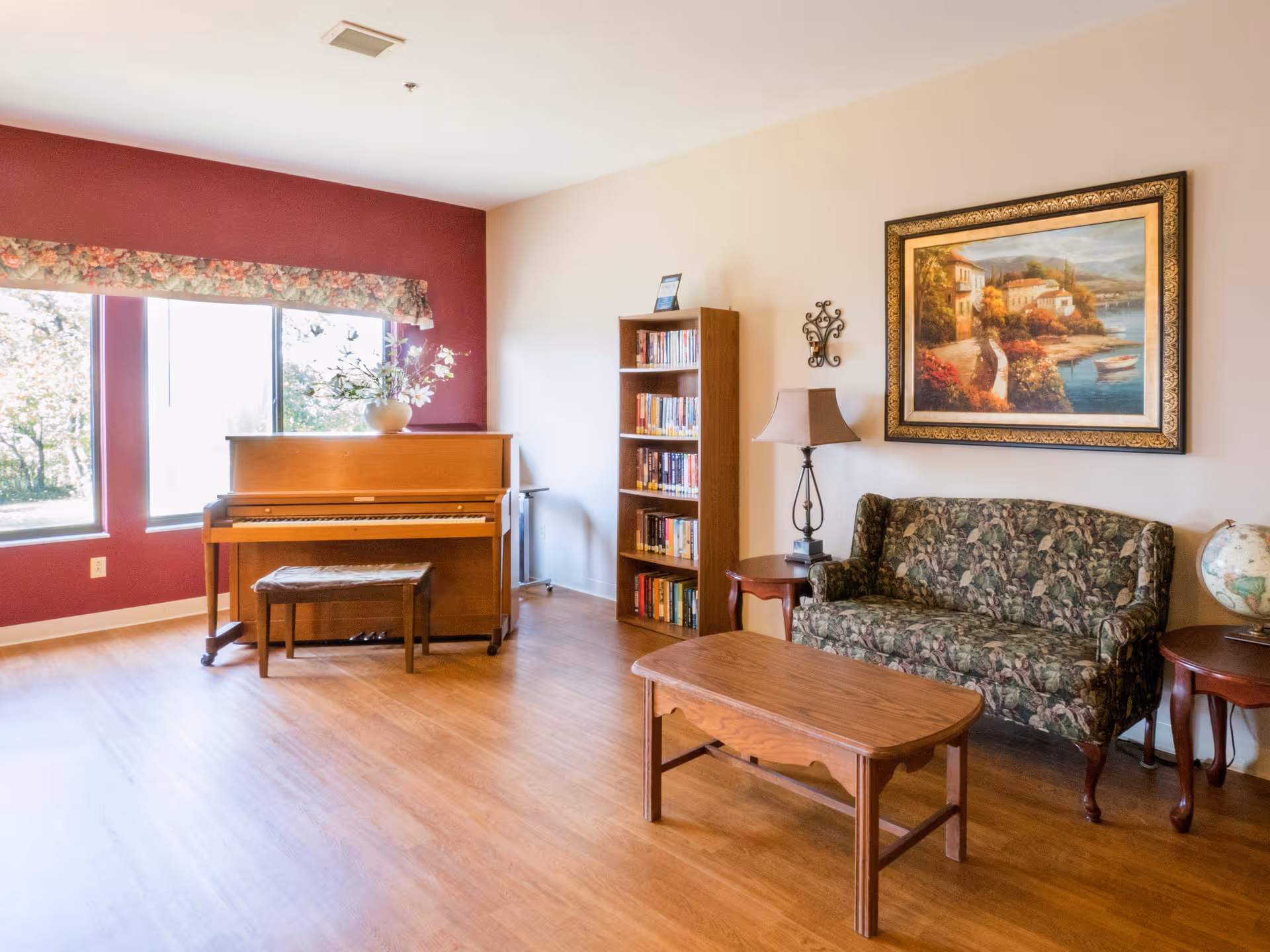 A cozy living room area with a wooden piano and matching bench near two large windows with floral valances. There is a wooden bookshelf filled with books, a floral patterned couch, a wooden coffee table, two side tables with a lamp and a globe, and a large framed painting of a coastal village on the wall.