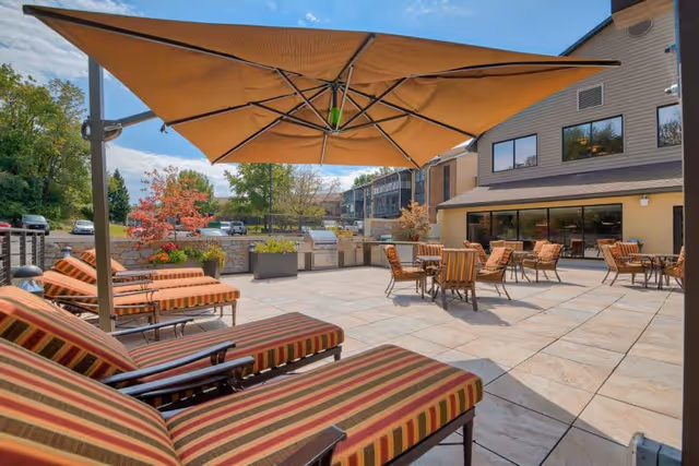 Outdoor patio area at Juniper Village at Bucks County featuring striped lounge chairs, several tables with chairs, a large beige umbrella, built-in grills, and potted plants under a clear blue sky.