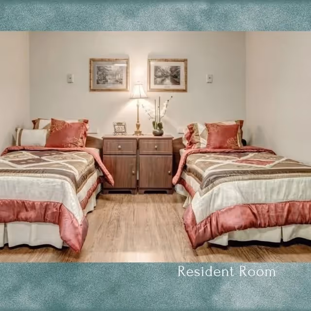 A resident room with two twin beds separated by a wooden nightstand with a lamp, a small plant, and a picture frame. Each bed has a brown, beige, and red patterned comforter with matching pillows. Two framed pictures hang on the wall above the nightstand.