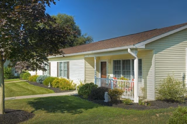 Single-story residential building with light yellow siding, a brown shingled roof, and a small front porch with white railing. The building is surrounded by well-maintained landscaping including bushes, a tree, and green grass under a clear blue sky.