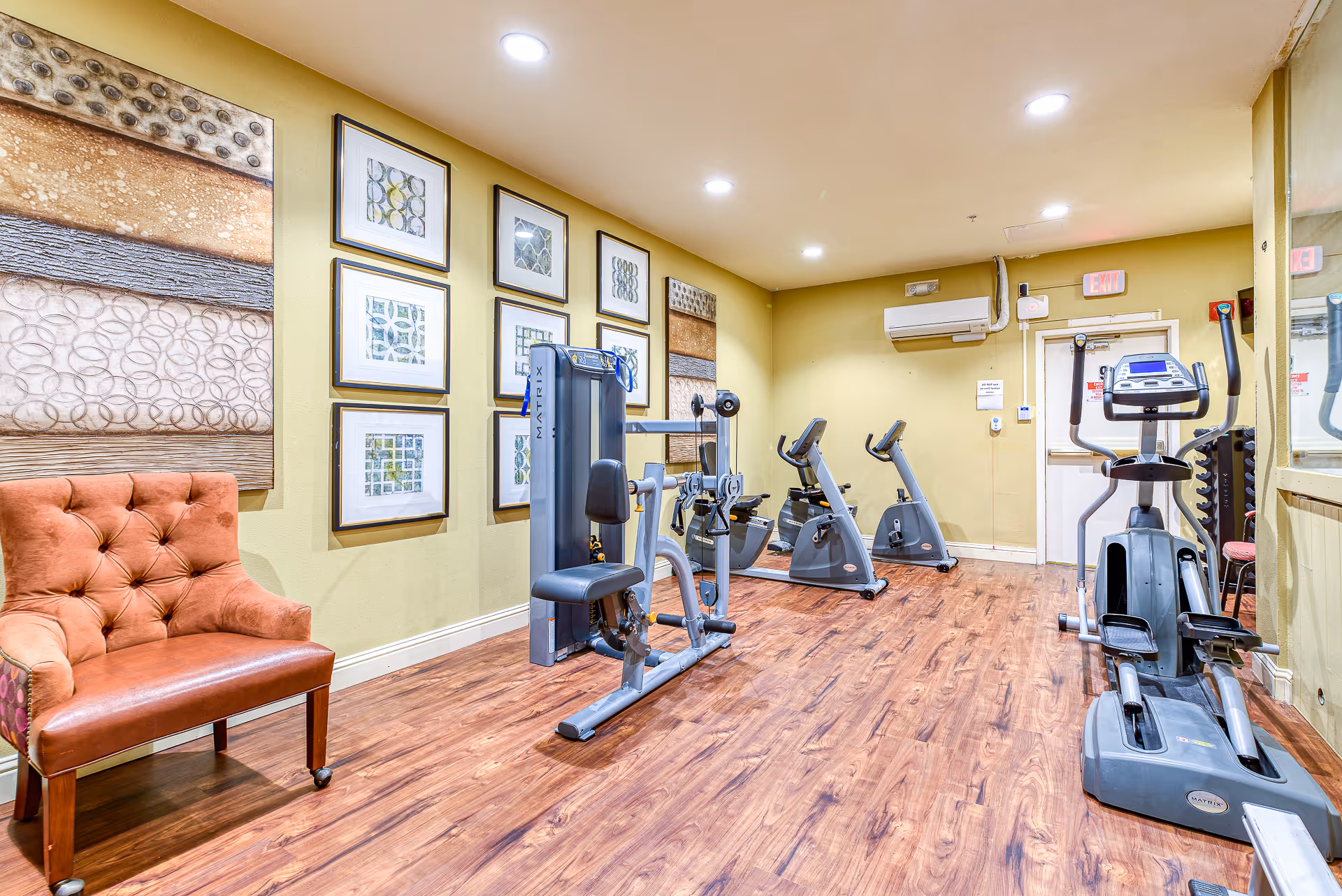A small fitness room with exercise equipment including a seated weight machine, two stationary bikes, and an elliptical trainer. The room has wooden flooring, yellow walls decorated with framed abstract art, and a brown upholstered chair in the corner. There is a large mirror on one wall and an air conditioning unit above the exit door.