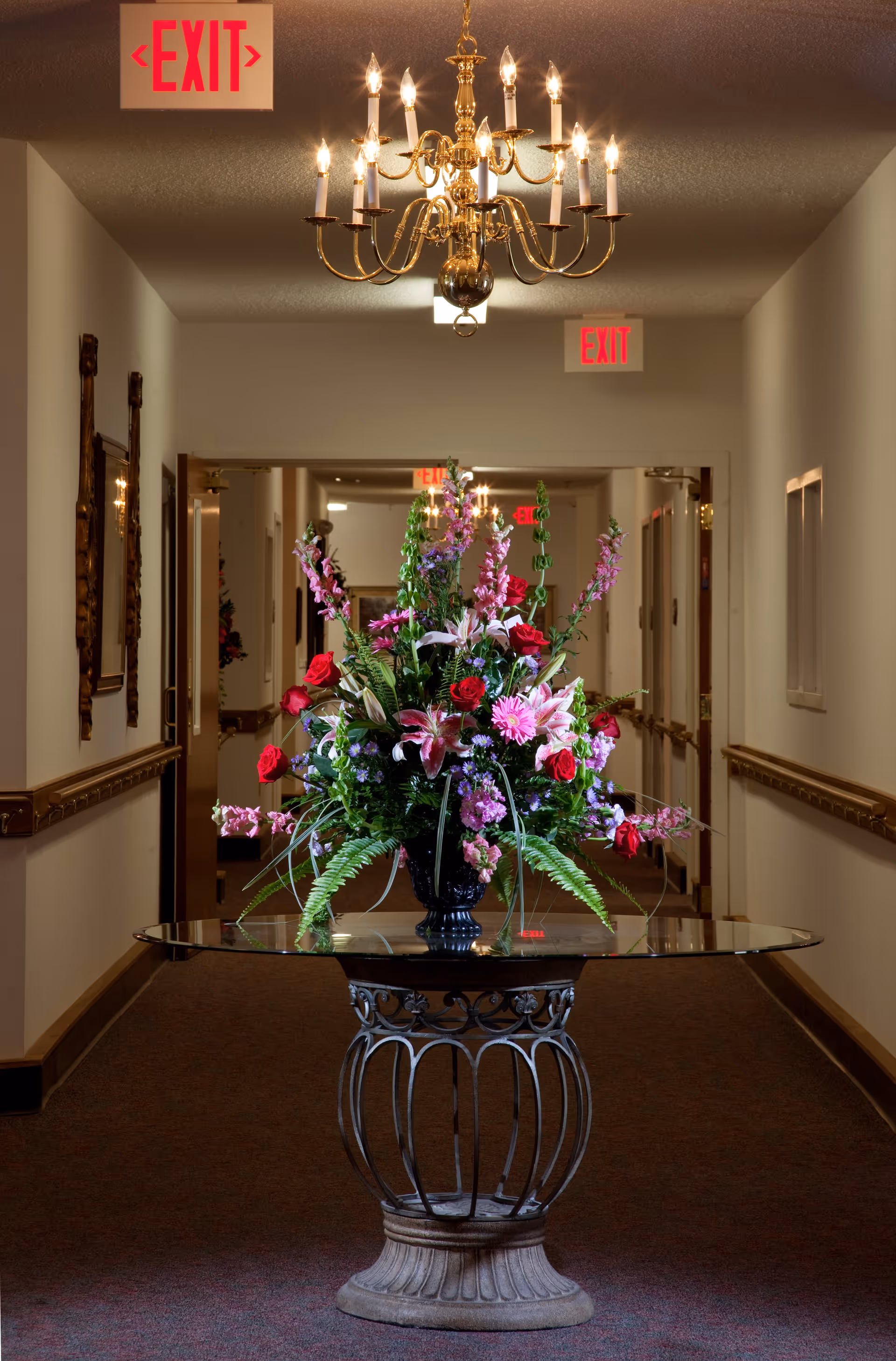 A hallway in a senior living facility with a decorative round glass table holding a large floral arrangement of red roses, pink lilies, and other flowers. The hallway has beige walls, carpeted floor, wooden handrails, and a gold chandelier hanging from the ceiling. Multiple illuminated red EXIT signs are visible in the background.