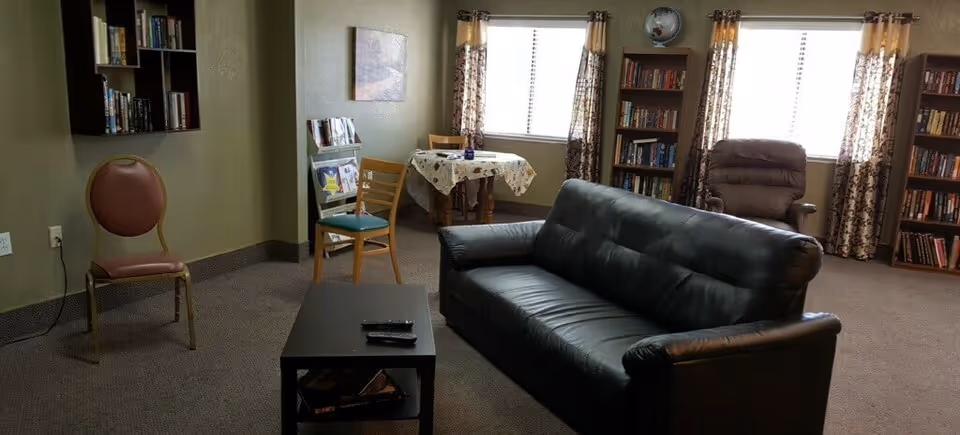 A cozy living room area with a black leather sofa, a brown recliner chair, and two wooden chairs. There is a small black coffee table with remote controls on it. The room has carpeted flooring, bookshelves filled with books, a small table covered with a floral tablecloth near the window, and curtains with a floral pattern. The walls are painted green and there is a globe on top of one bookshelf.