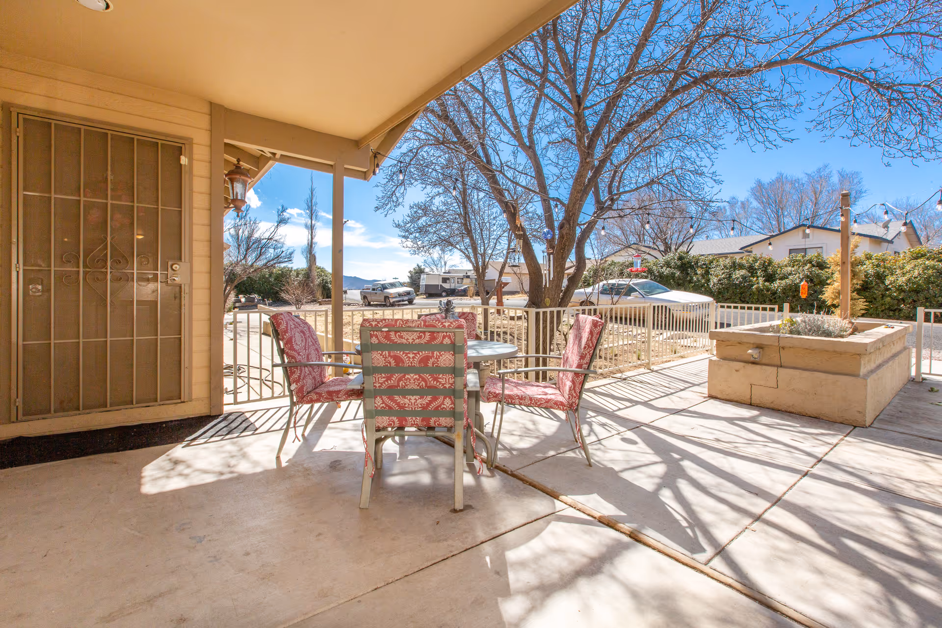 Outdoor patio area with a round glass table and four metal chairs with red patterned cushions. The patio is covered and has a concrete floor. In the background, there are leafless trees, a parked car, and a residential neighborhood under a clear blue sky.