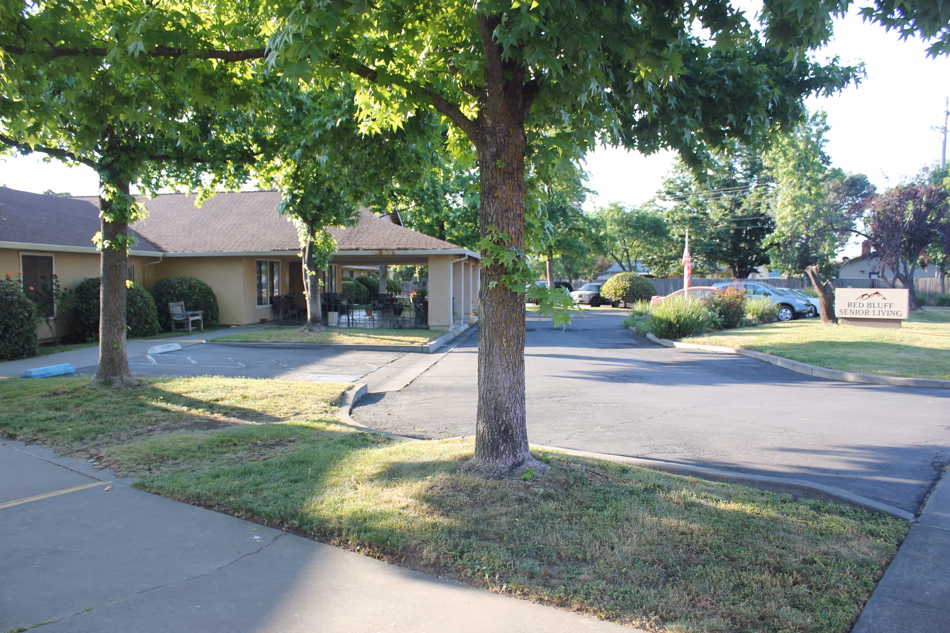 Exterior view of Red Bluff Senior Living facility showing a parking area, trees, and a building with a covered patio. A sign with the facility's name is visible on the right side near the parking lot.