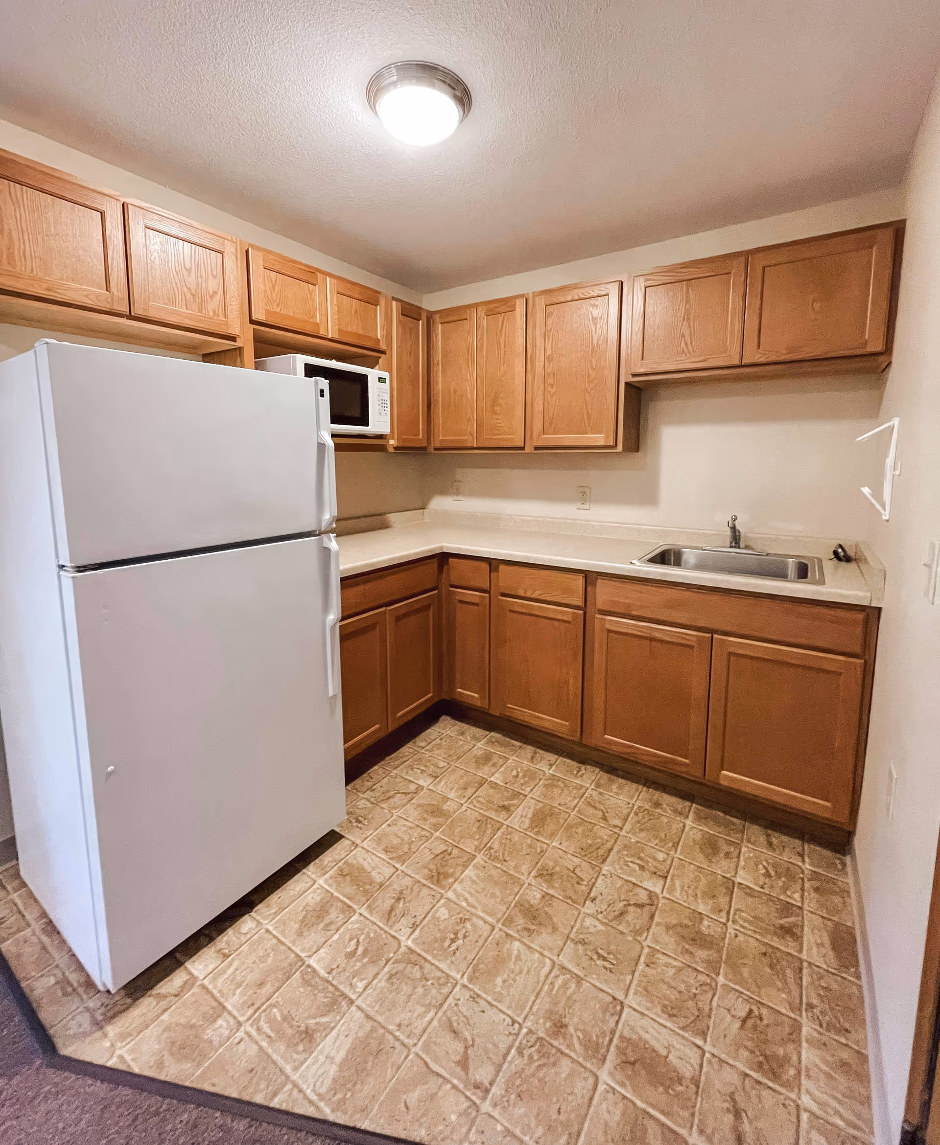Small kitchen area with wooden cabinets, a white refrigerator, a microwave, a stainless steel sink, and beige tiled flooring.