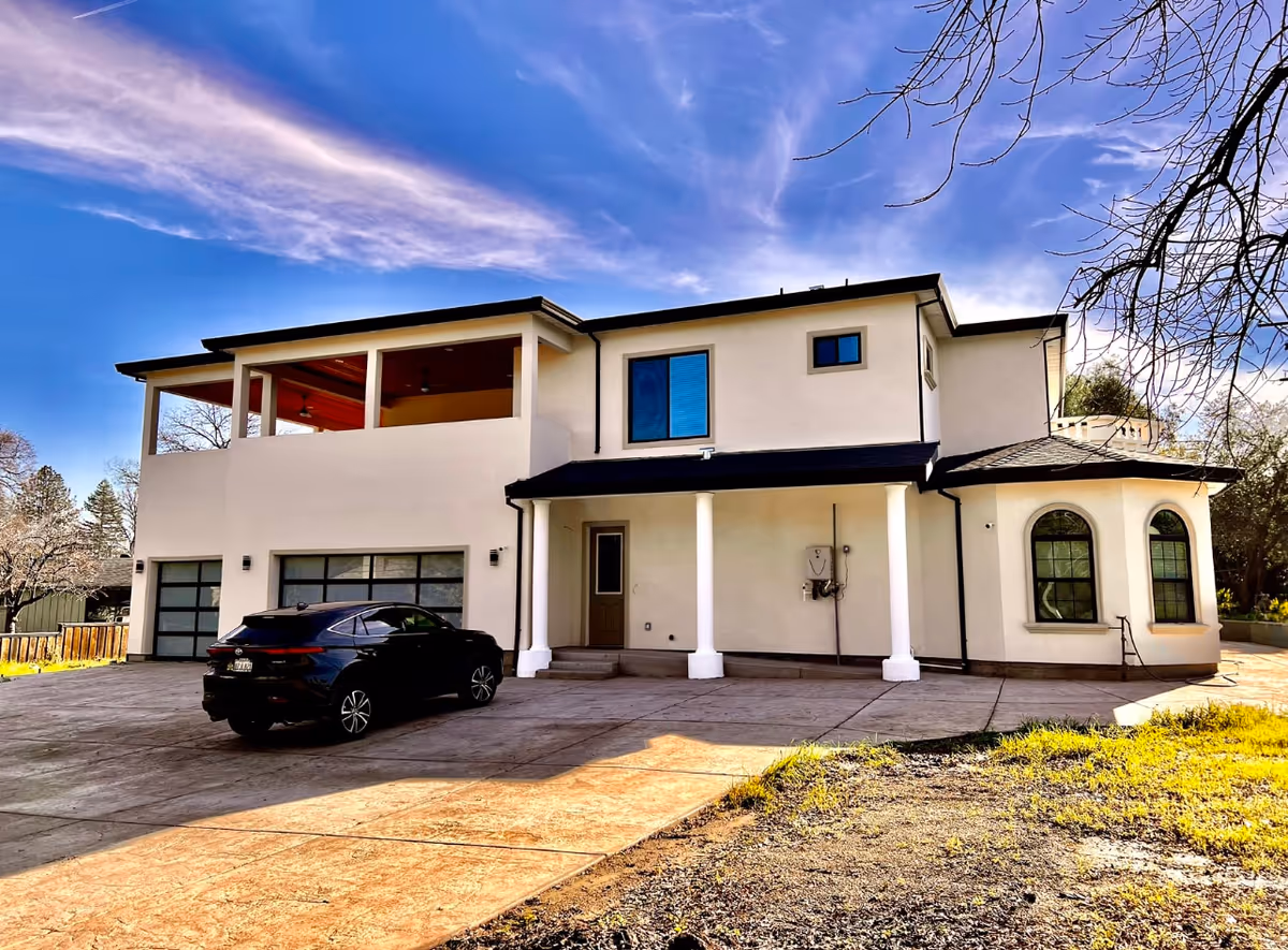 Exterior view of a two-story white building with a covered porch supported by columns, large windows, and a black car parked in the driveway under a blue sky with wispy clouds.