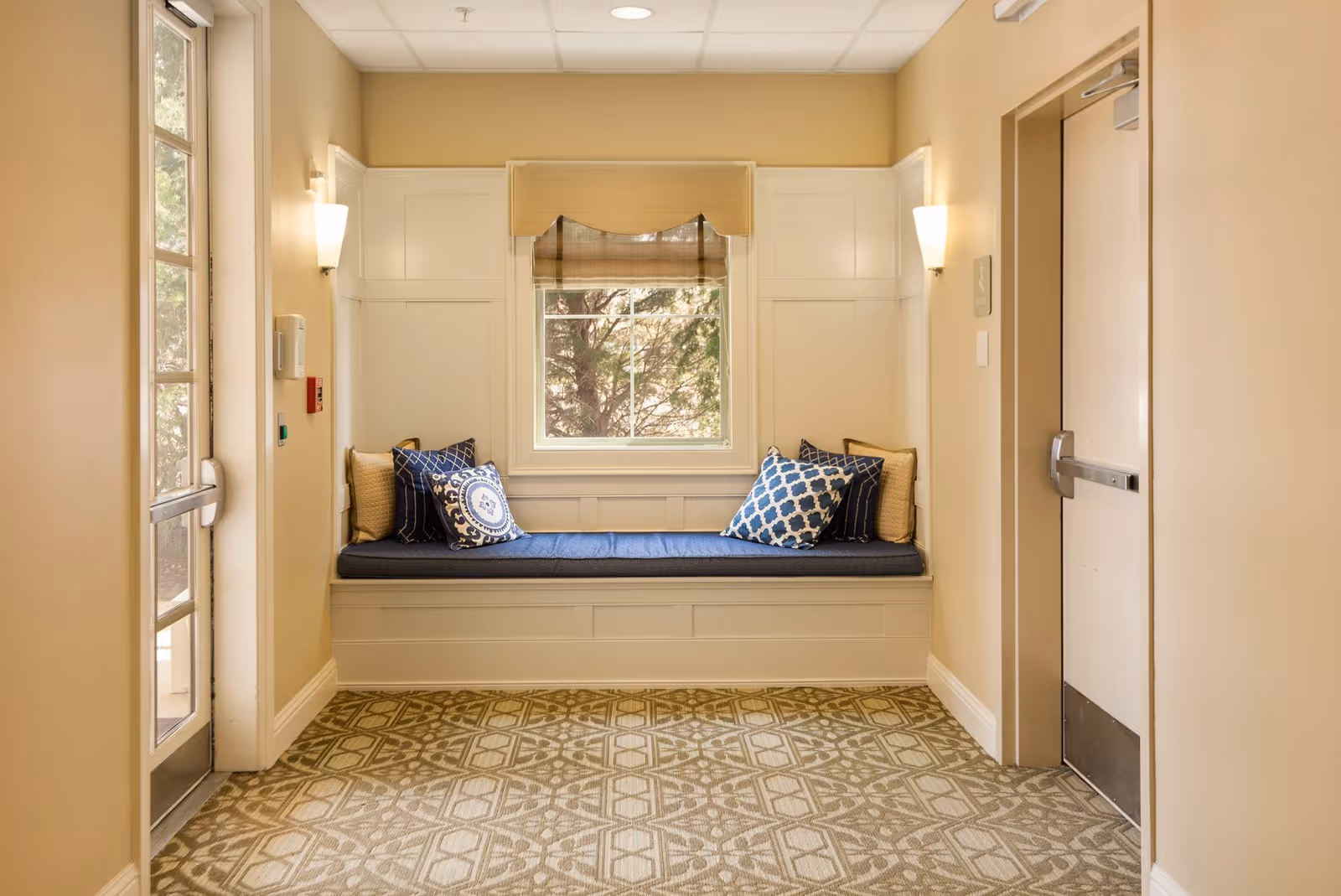 A cozy window nook with a cushioned bench seat adorned with several decorative pillows in blue and beige tones. The nook is framed by white paneling and a window with a beige roman shade, allowing natural light to filter in. The surrounding walls are painted a warm beige, with two wall sconces providing additional lighting. To the left is a glass door, and to the right is a closed door with a metal push bar. The floor is covered with a patterned carpet in neutral colors.