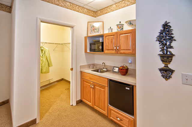 Small kitchenette with wooden cabinets, sink, mini-fridge and microwave next to a walk-in closet.