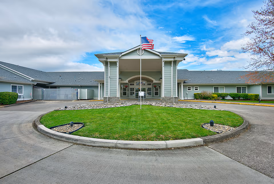 Front exterior view of Spring Valley Assisted Living facility with a circular driveway, a green lawn in the center, an American flag on a flagpole, and a cloudy blue sky above.