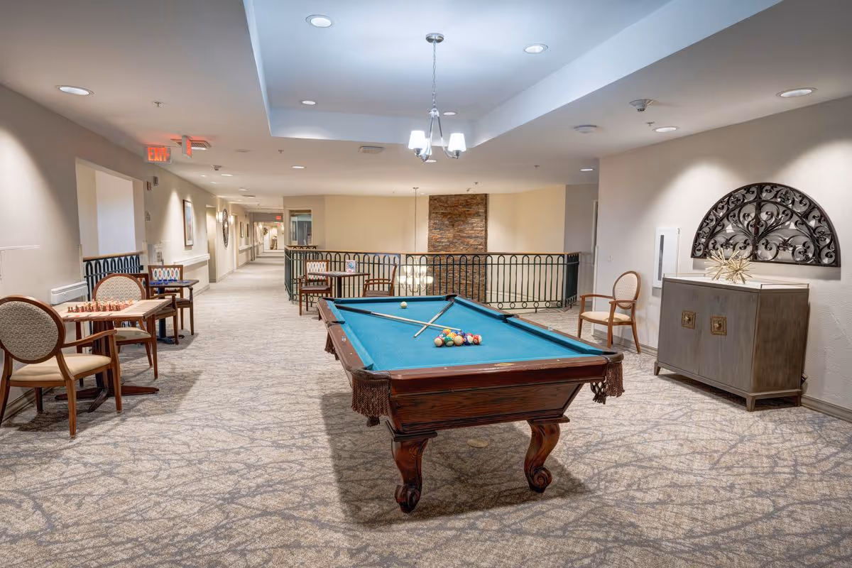 A spacious indoor common area featuring a pool table with billiard balls and cues set up, several small tables with chairs, one table with a chessboard, and decorative wall art. The area is well-lit with ceiling lights and has a carpeted floor. A railing overlooks a lower level, and a hallway extends into the distance.