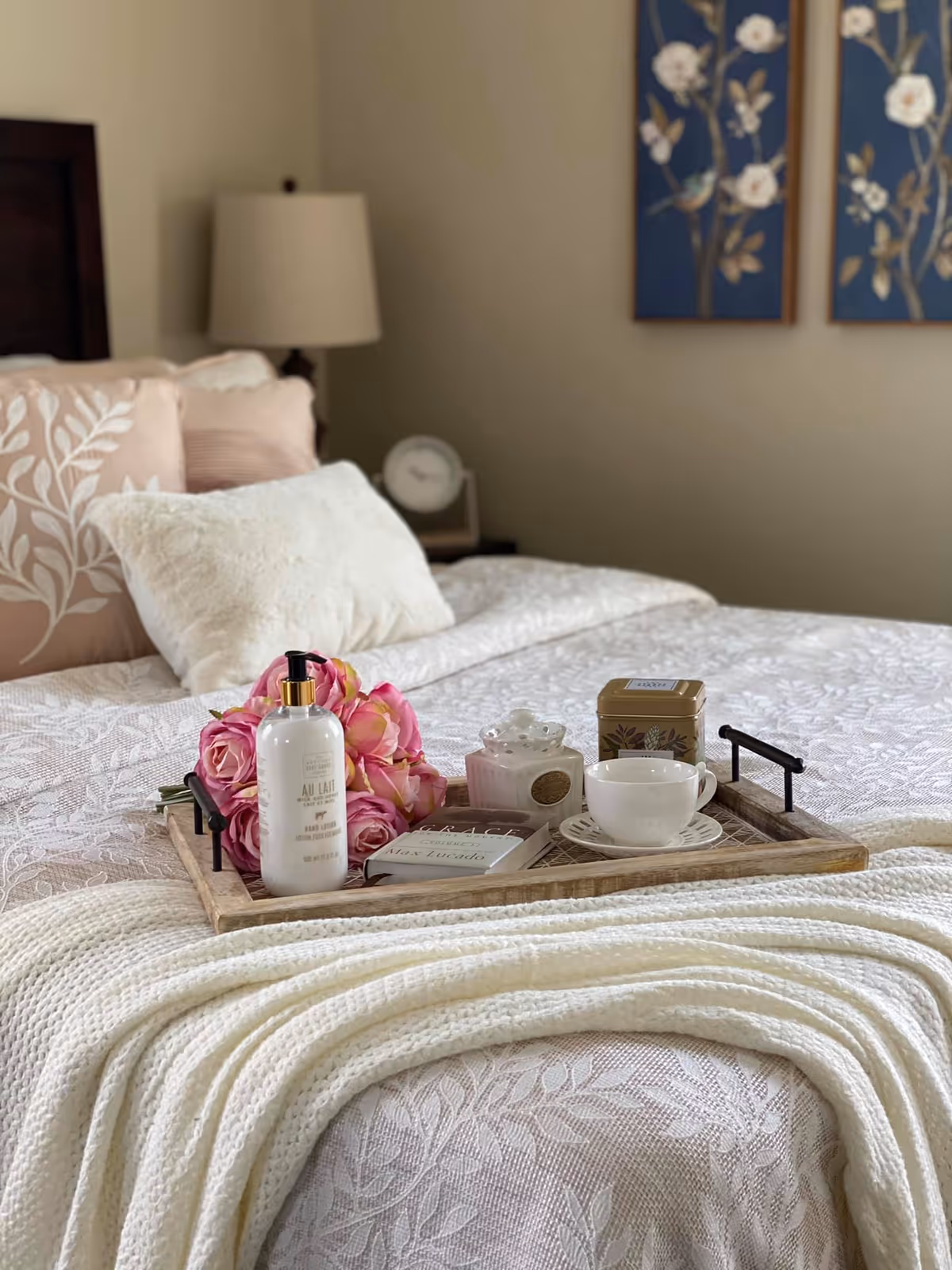 A neatly made bed in a softly lit bedroom with decorative pillows and a wooden tray holding flowers, a teacup, and toiletries.
