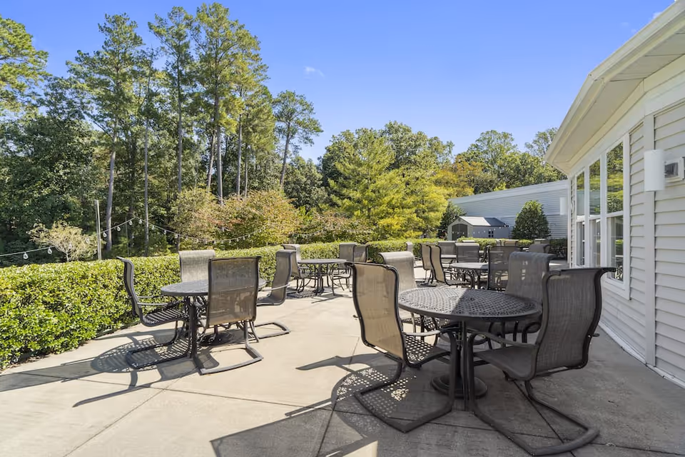 Outdoor patio area with several round metal tables and mesh chairs arranged on a concrete surface. The patio is bordered by green hedges and surrounded by tall trees under a clear blue sky. The side of a light-colored building with large windows is visible on the right.
