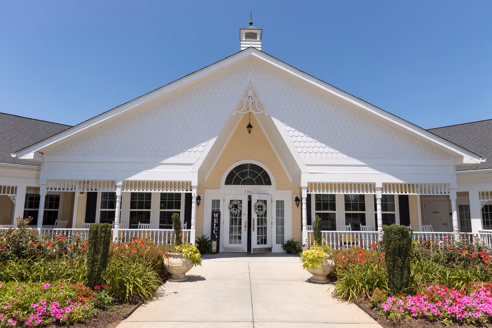 Front exterior view of Cadence at Clemmons facility with a white building featuring a peaked roof, double glass doors with wreaths, a welcome sign, and a well-maintained garden with colorful flowers and shrubs in front.