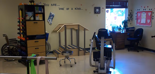 A therapy or rehabilitation room with exercise equipment including a stationary bike, parallel bars for walking practice, a set of stairs for physical therapy, a rack of colorful dumbbells, a wheelchair, and two desks with chairs near a window. The wall has a motivational message that reads 'You are... one of a kind.'