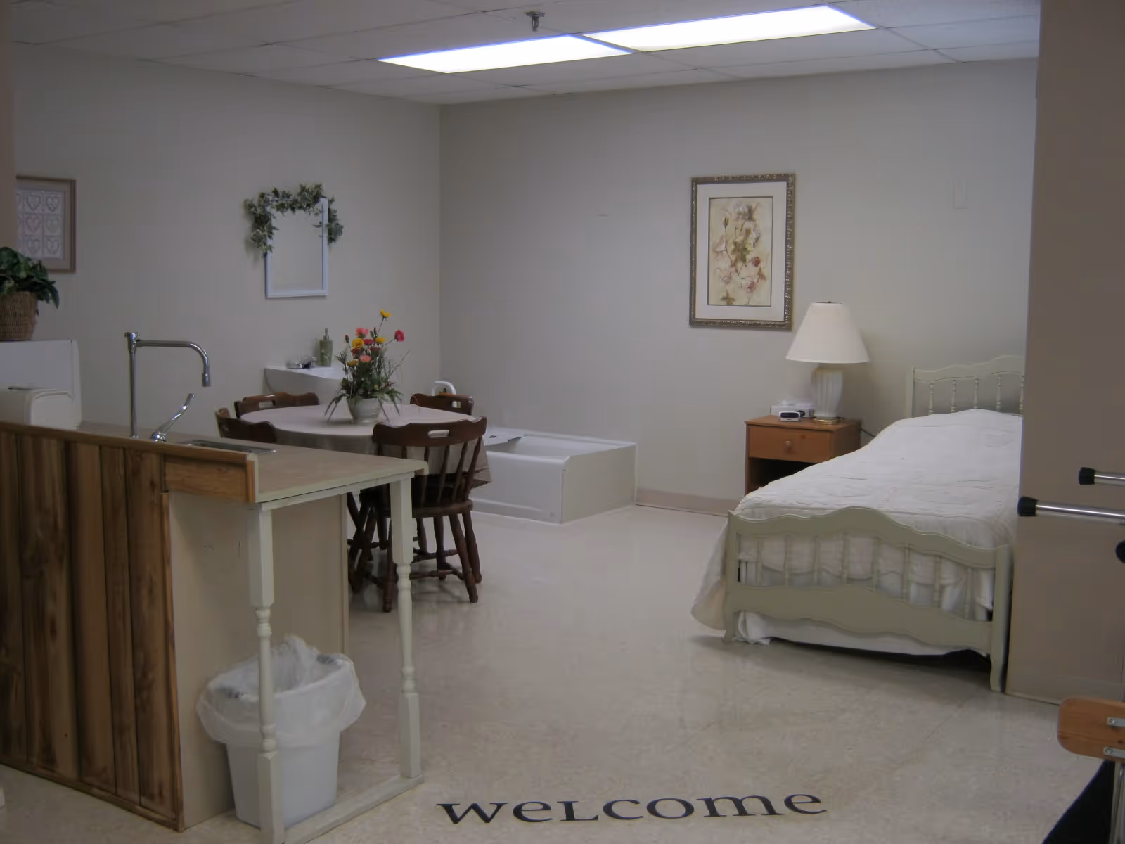Interior view of a senior living facility room featuring a single bed with white bedding, a wooden nightstand with a lamp, a small dining table with four chairs and a flower arrangement, a small sink area with a mirror and decorative greenery, and a partial view of a kitchenette with a faucet and trash can. The floor has the word 'welcome' written on it.