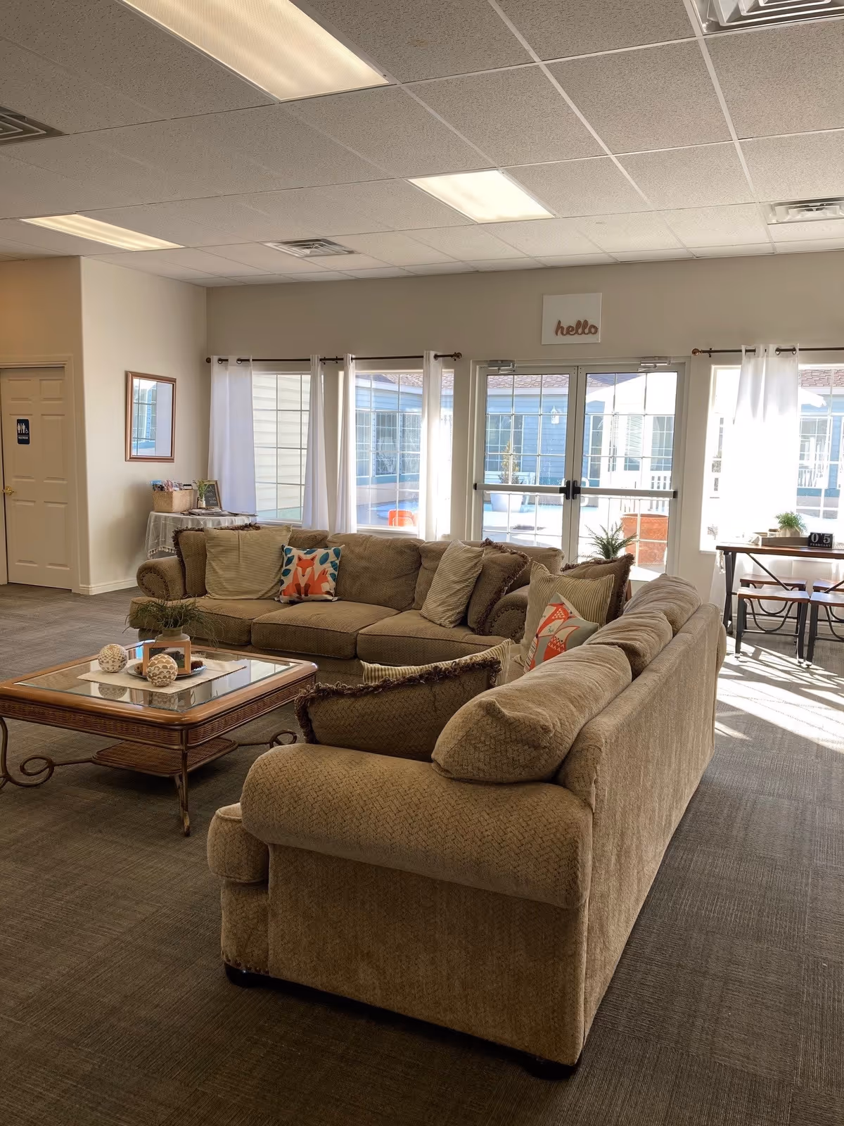A bright and cozy living room area with two beige sofas arranged around a wooden coffee table with decorative items. Large windows and glass doors let in natural light, with white curtains framing the windows. A small table with chairs is visible near the windows, and a sign above the doors reads 'hello'. The room has a carpeted floor and a drop ceiling with fluorescent lights.