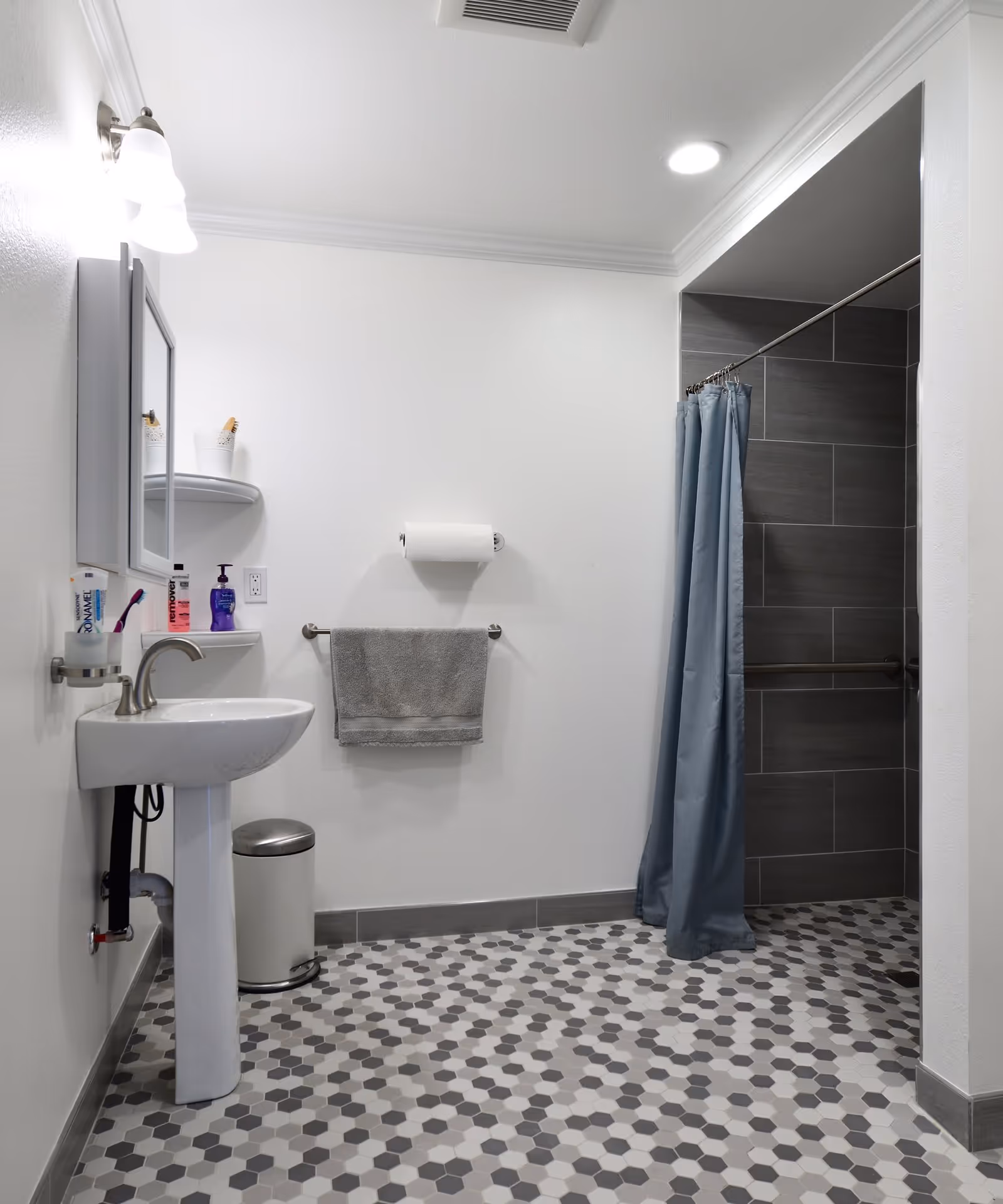 Bright accessible bathroom with a pedestal sink, towel and toilet paper on the wall, a tiled walk-in shower with a blue curtain, and gray hexagonal floor tiles.