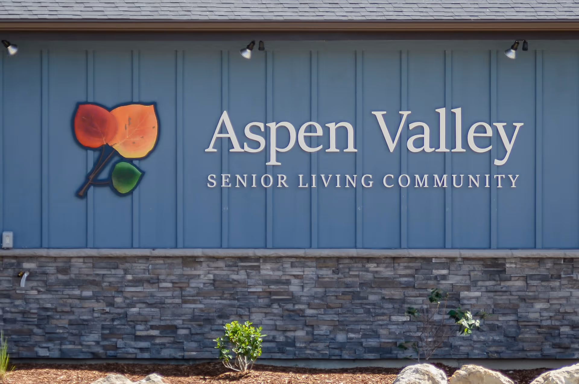 Exterior wall of Aspen Valley Senior Living Community with a blue panel background and stone facade below. There is a decorative sign featuring three colorful leaves and the text 'Aspen Valley Senior Living Community'. Small plants and rocks are visible at the base of the wall.