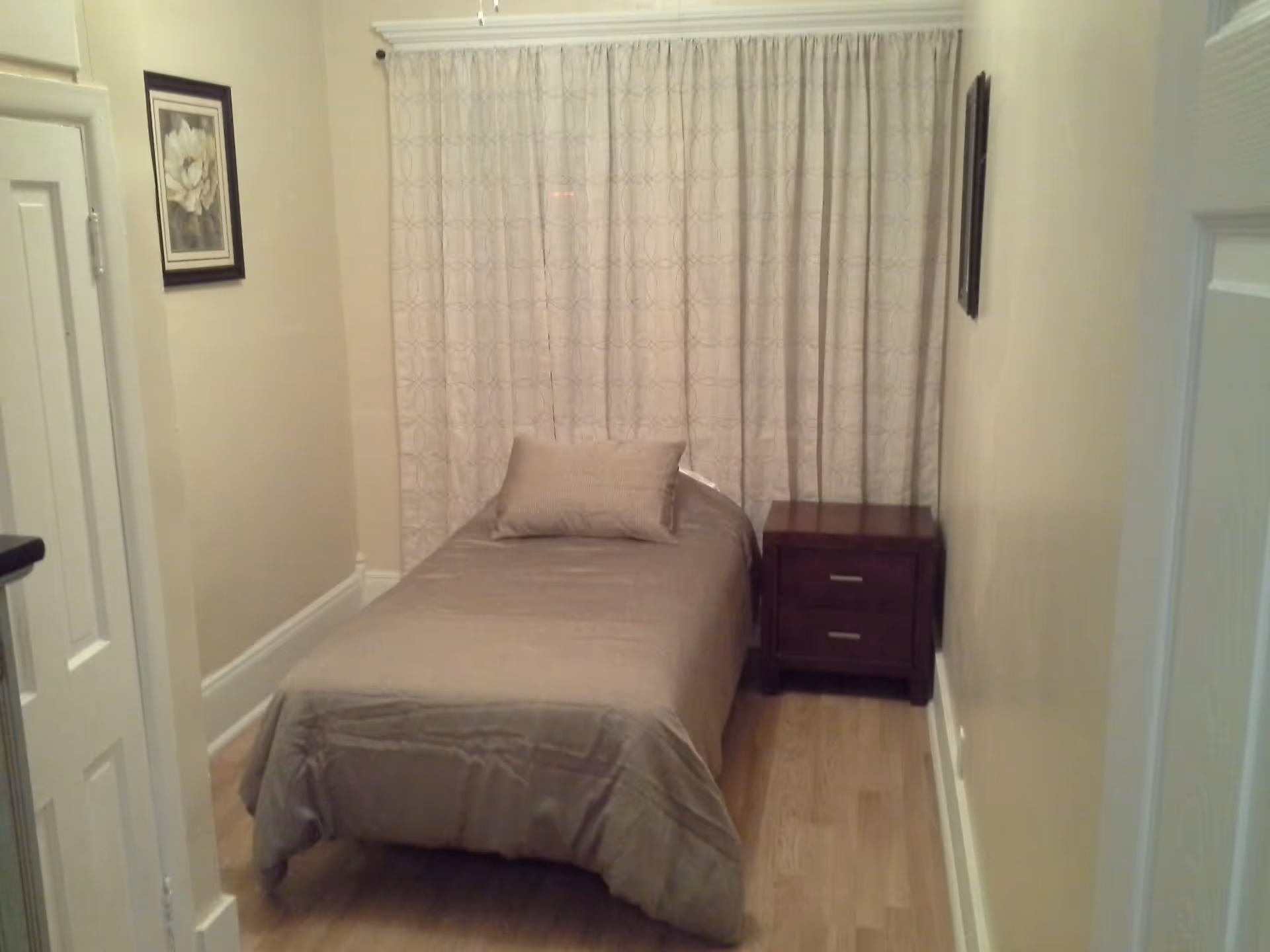 Small bedroom with a single bed, beige bedding, a wooden nightstand, and patterned curtains.