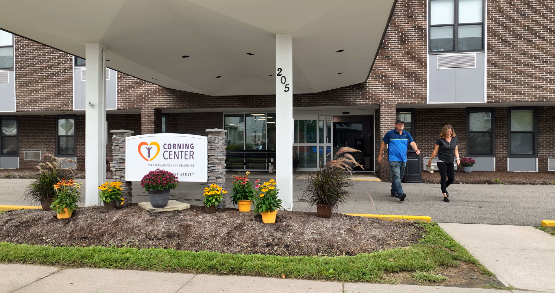 Entrance of Corning Center for Rehabilitation and Nursing with a covered drop-off area. Two people are walking away from the entrance. There are flower pots with colorful flowers arranged around a sign that displays the facility's name and logo.