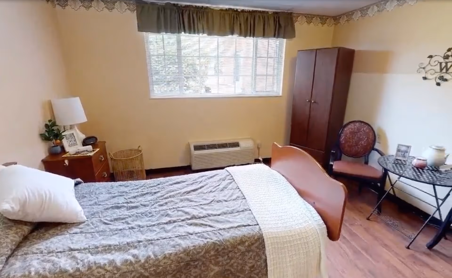 A cozy bedroom with a single bed covered in a patterned gray bedspread and a white pillow. Next to the bed is a wooden nightstand with a lamp, a small plant, and a framed photo. A window with blinds and a valance lets in natural light. Against the far wall is a wooden wardrobe, a round table with a decorative item and a candle, and a cushioned chair with a patterned backrest. The room has wooden flooring and light-colored walls with a decorative border near the ceiling.
