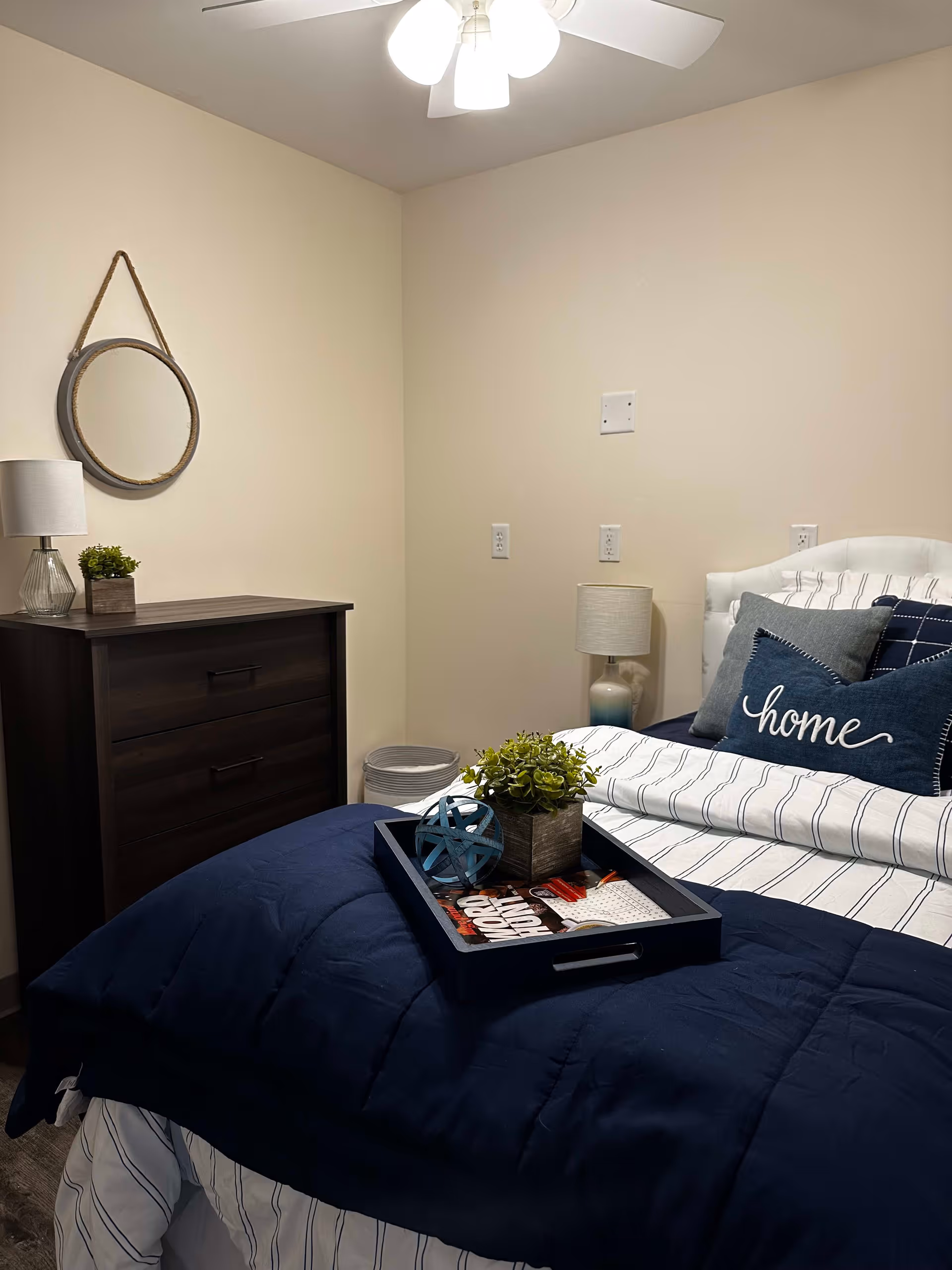 A cozy bedroom with a neatly made bed featuring navy blue and white striped bedding and decorative pillows, including one with the word 'home'. On the bed is a tray holding a small plant and decorative items. Next to the bed is a nightstand with a lamp, and across from it is a dark wooden dresser with a small plant and a lamp. A round mirror hangs on the wall above the dresser. The room has beige walls and a ceiling fan with lights.