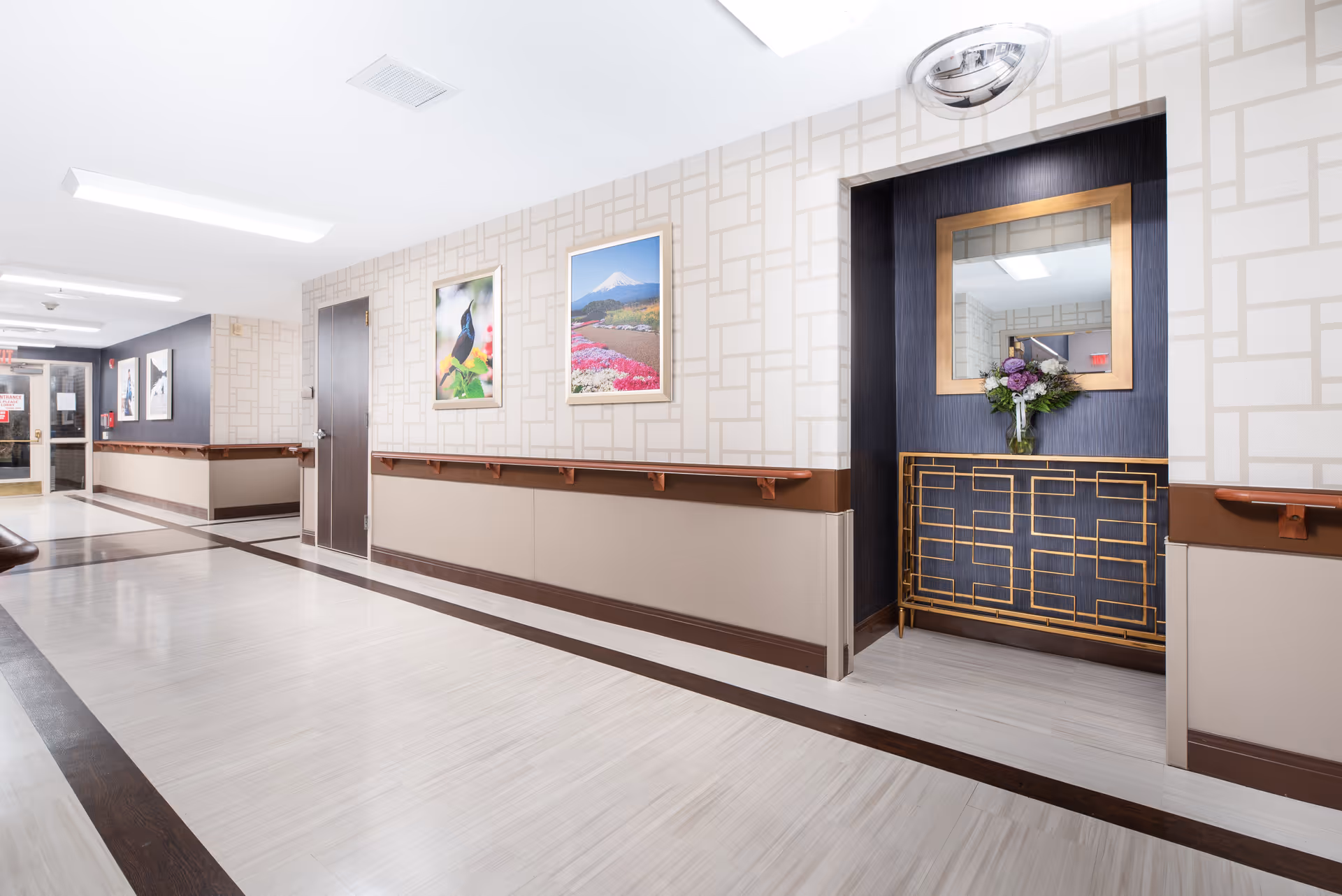 A clean and well-lit hallway in a senior living facility with light-colored flooring and walls decorated with framed nature photographs. There is a decorative console table with a mirror and a vase of flowers in a recessed area on the right side. Handrails run along both sides of the hallway.