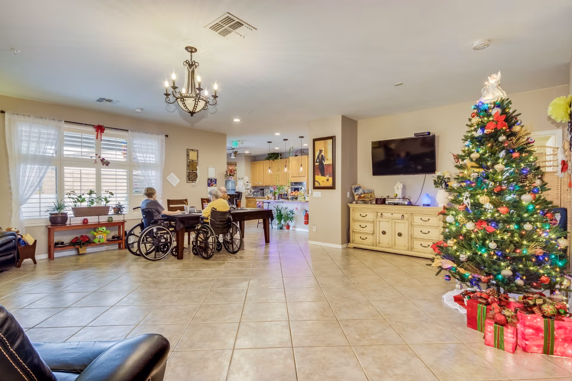 A spacious, well-lit common area in an assisted living facility decorated for Christmas with a large decorated Christmas tree and wrapped presents. Two elderly individuals in wheelchairs are seated at a wooden table near a window with sheer curtains and potted plants. The room features tiled flooring, a chandelier, a wall-mounted TV above a cream-colored cabinet, and an open kitchen area in the background.