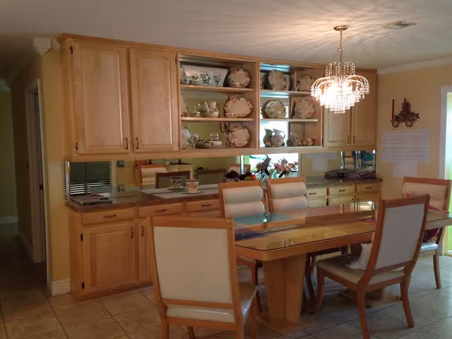 A dining area with a wooden table and six cushioned chairs. Above the table hangs a decorative chandelier. Behind the table is a wooden cabinet with shelves displaying a collection of floral-patterned plates and teapots. The room has tiled flooring and light yellow walls.