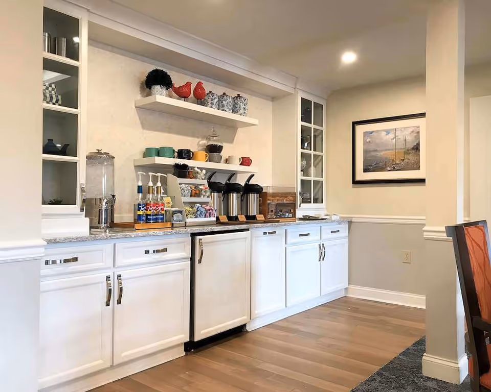 A bright and clean kitchenette area with white cabinets and shelves. The countertop holds coffee dispensers, syrup bottles, a water dispenser, and a variety of mugs and cups on shelves above. A framed painting hangs on the wall to the right, and part of a chair with an orange cushion is visible on the right side.