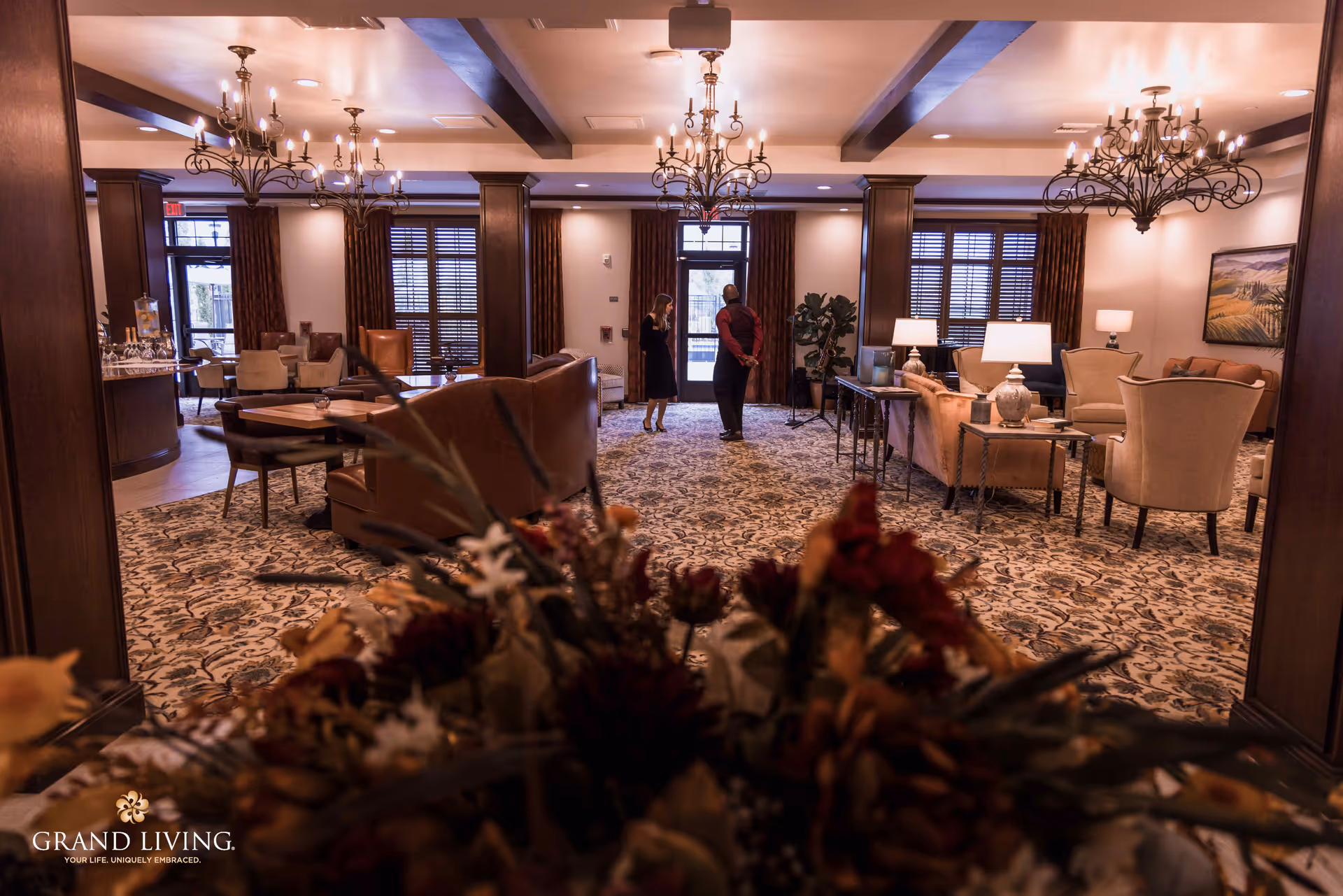 Warmly lit senior living community lounge with chandeliers, multiple seating areas, and two people standing near the entrance.