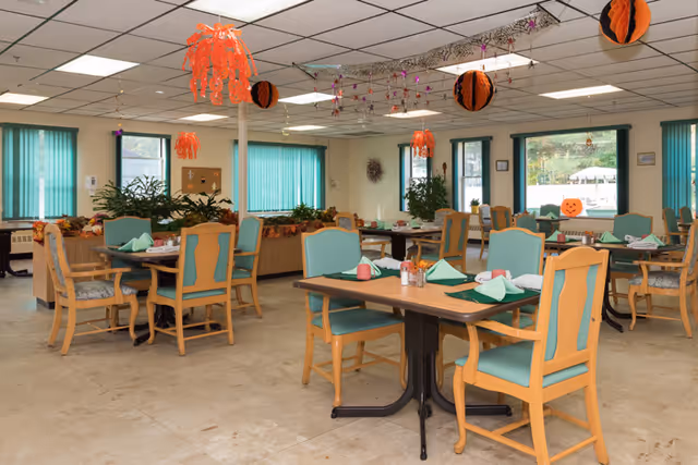 A dining room in a senior living facility decorated with Halloween-themed hanging decorations. The room has wooden tables with green cushioned chairs, each table set with green napkins and place settings. Large windows with teal vertical blinds let in natural light, and there are plants and seasonal decorations around the room.