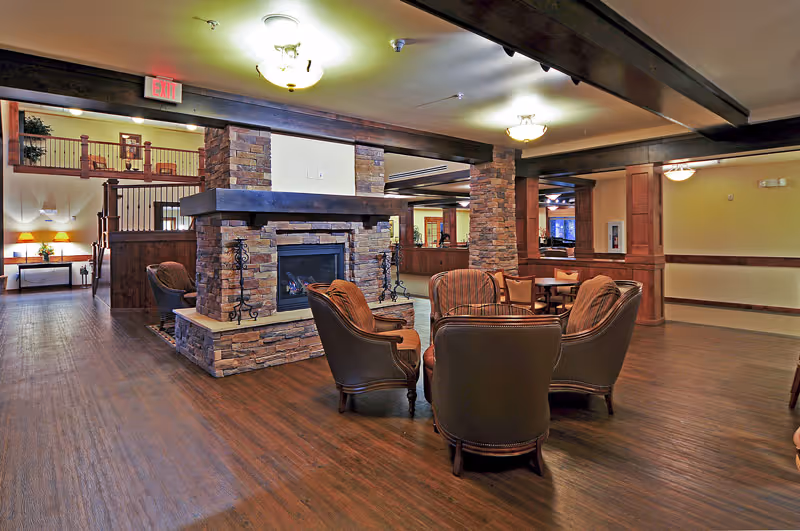 Spacious senior living common area with leather chairs arranged around a central stone fireplace under wood-beamed ceilings.