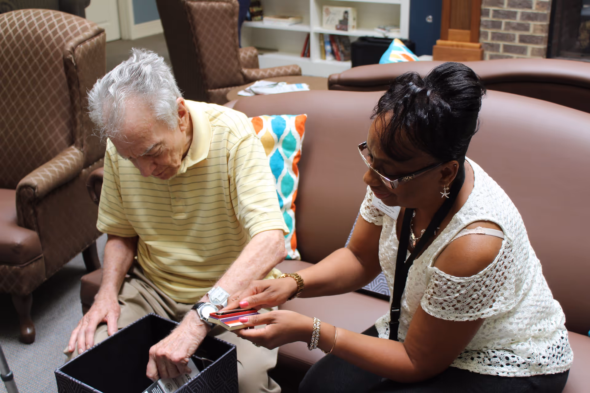 An elderly man sitting on a couch next to a woman who is showing him a set of color swatches. They are in a cozy living room with brown chairs, a bookshelf, and a fireplace in the background.