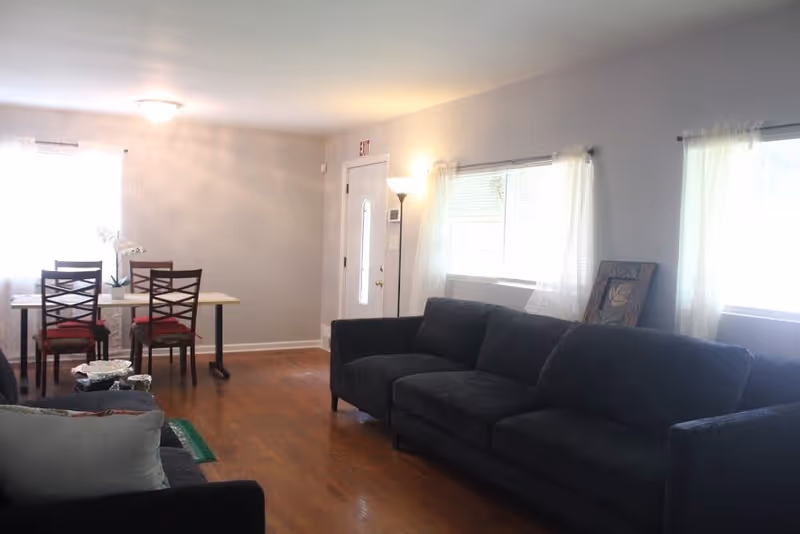 A bright living room with a large dark gray sofa along the right wall and a smaller sofa with cushions in the foreground. There are two windows with sheer white curtains letting in natural light. In the background, a dining area with a rectangular table and four chairs with red cushions is visible. The room has wooden flooring and light-colored walls. A floor lamp is near the entrance door, which has an exit sign above it.