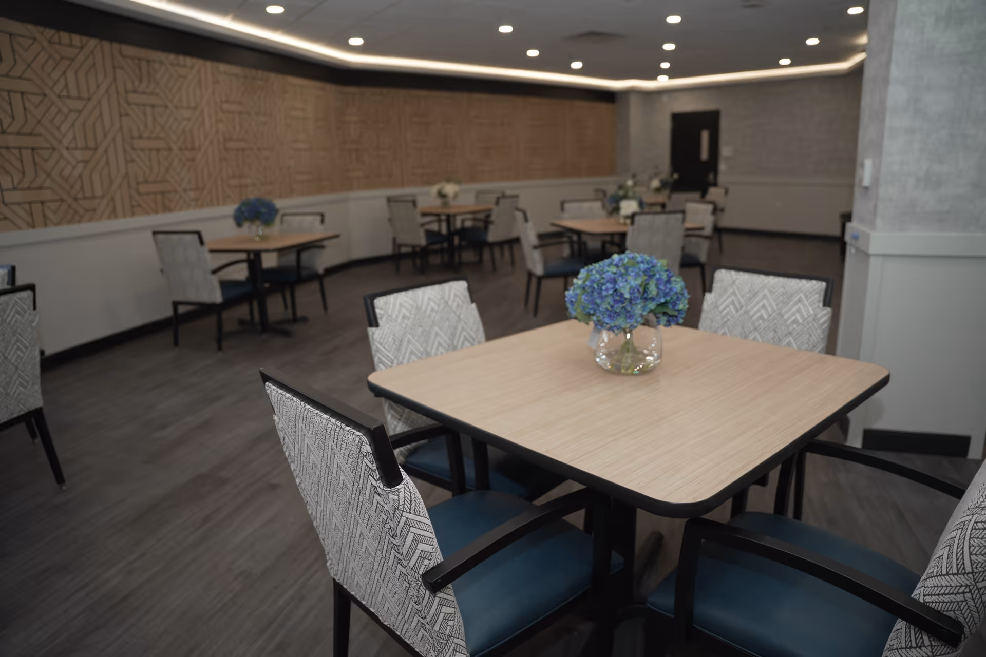 A dining room with several square wooden tables, each surrounded by four cushioned chairs with patterned backs and blue seats. Each table has a small glass vase with blue flowers. The room has a modern design with a patterned wall, gray flooring, and recessed ceiling lights.