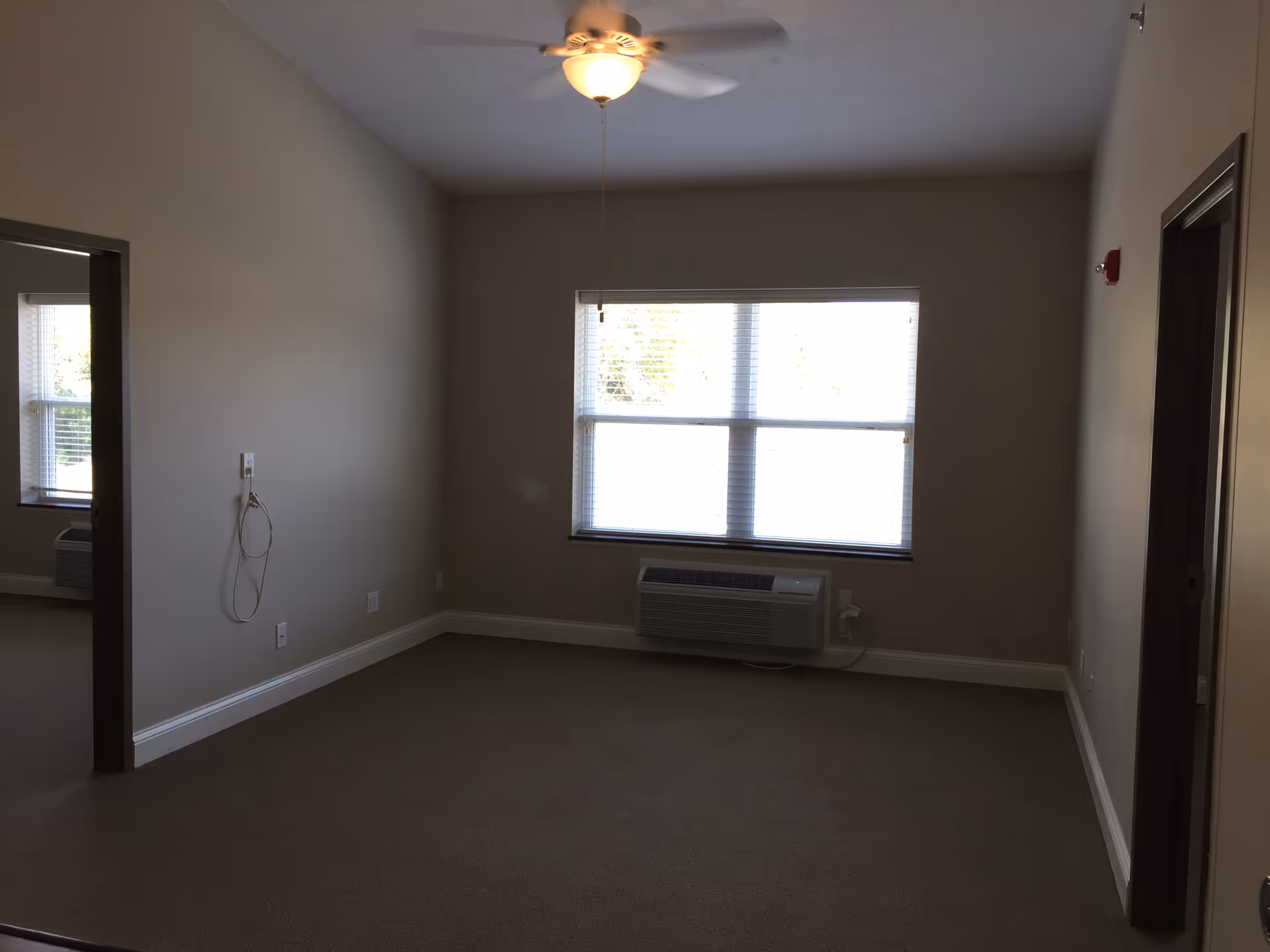 Empty apartment living room with beige walls, a ceiling fan, a large window and an air conditioning unit beneath it.