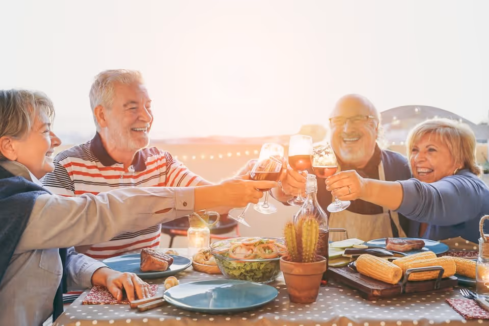 Four elderly people sitting around a table outdoors, smiling and clinking their wine glasses together in a toast. The table is set with plates of food including corn on the cob, salad, and steak. There is a small potted cactus on the table and string lights in the background.