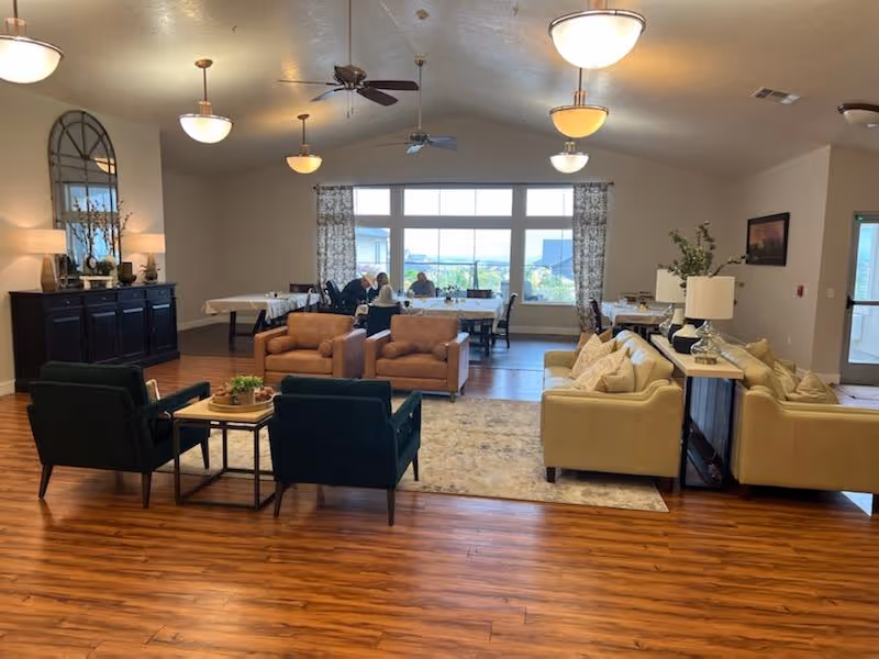 A spacious and well-lit common area in an assisted living facility featuring comfortable seating including two beige sofas, two brown armchairs, and two dark green chairs arranged around small tables on a rug. In the background, there are dining tables with people seated near large windows with patterned curtains letting in natural light. The room has wooden flooring, ceiling fans, and multiple ceiling lights. A dark wooden sideboard with a large mirror and decorative items is visible on the left side.