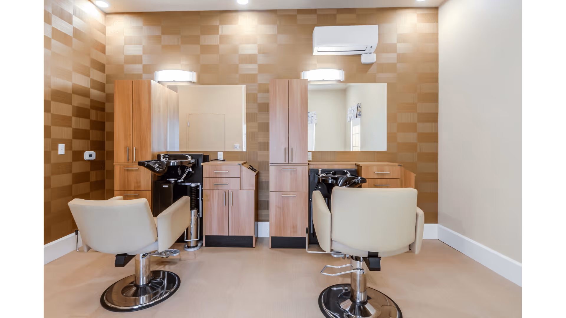 Interior view of a salon area with two beige salon chairs facing mirrors mounted on a wall with a checkered pattern. Each station has wooden cabinets and black hair washing sinks. The room has a clean and modern design with neutral tones.