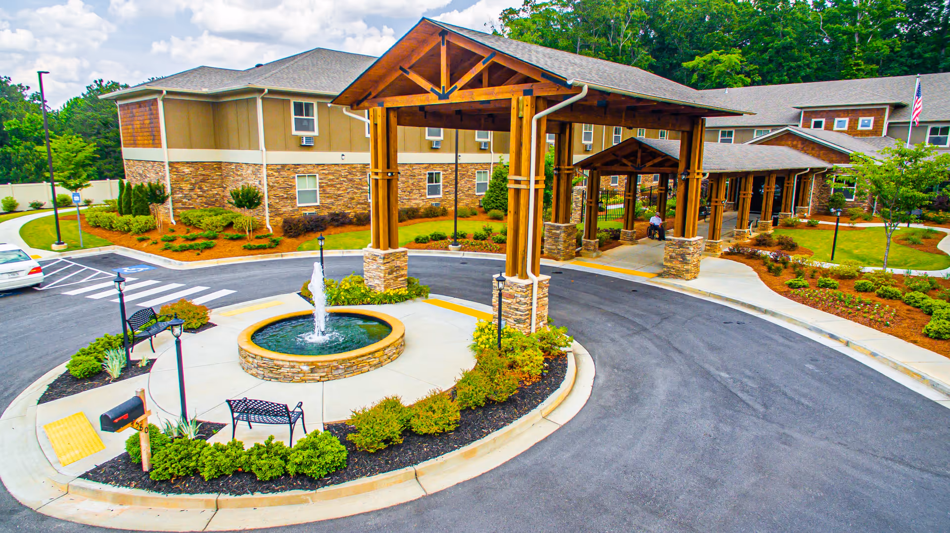 Entrance area of Oaks at Towne Lake senior living facility featuring a circular driveway with a central fountain, wooden covered walkways supported by stone pillars, landscaped greenery, benches, and a person in a wheelchair near the entrance.