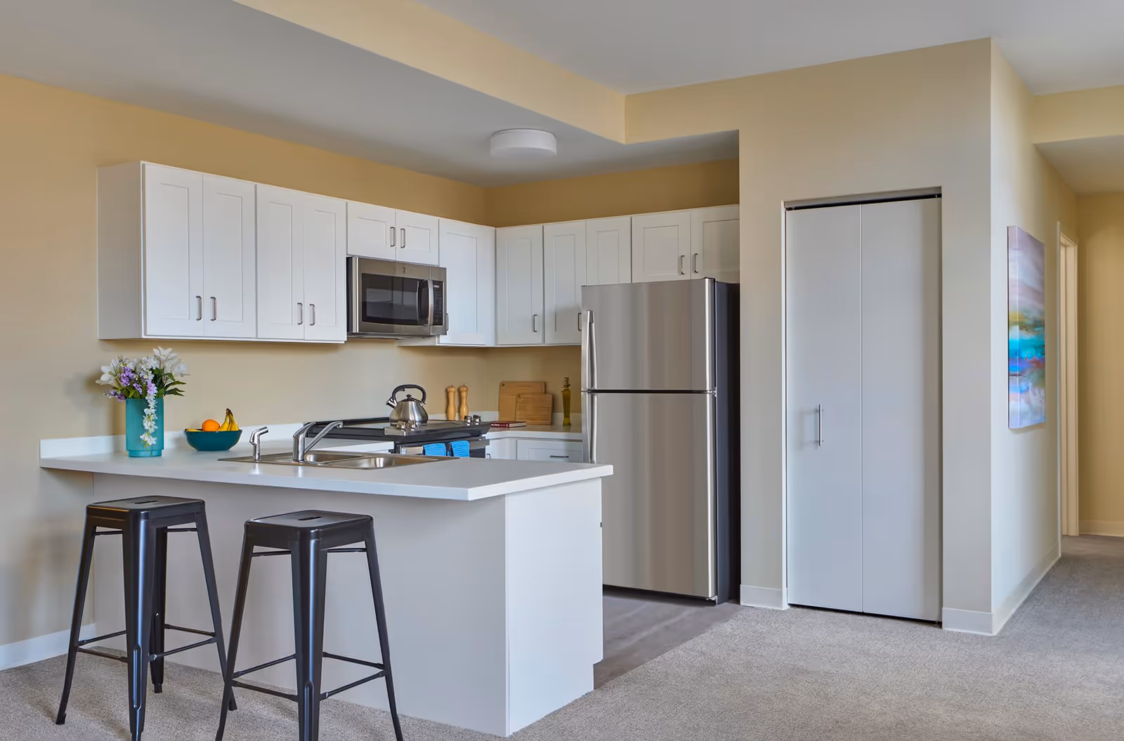 Modern kitchen area with white cabinets, stainless steel refrigerator and microwave, a white countertop with a sink, two black bar stools, a bowl of fruit, and a vase with flowers. The kitchen opens into a carpeted hallway with a closed white closet door and a colorful painting on the wall.