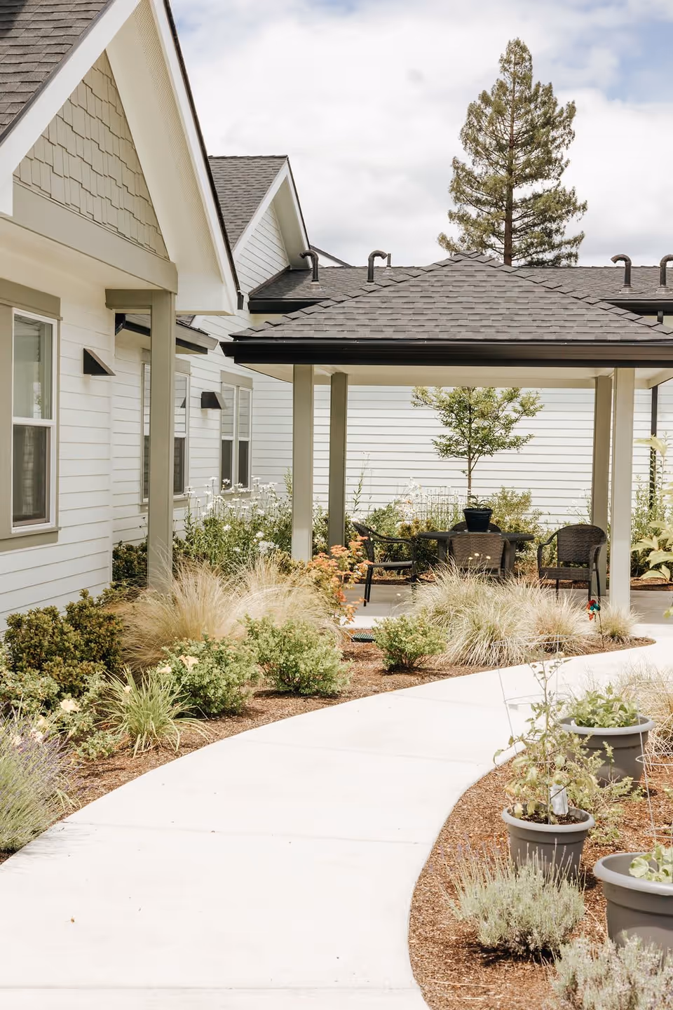 A curved concrete path winds through landscaped gardens to a covered patio with chairs between light-colored residential buildings.