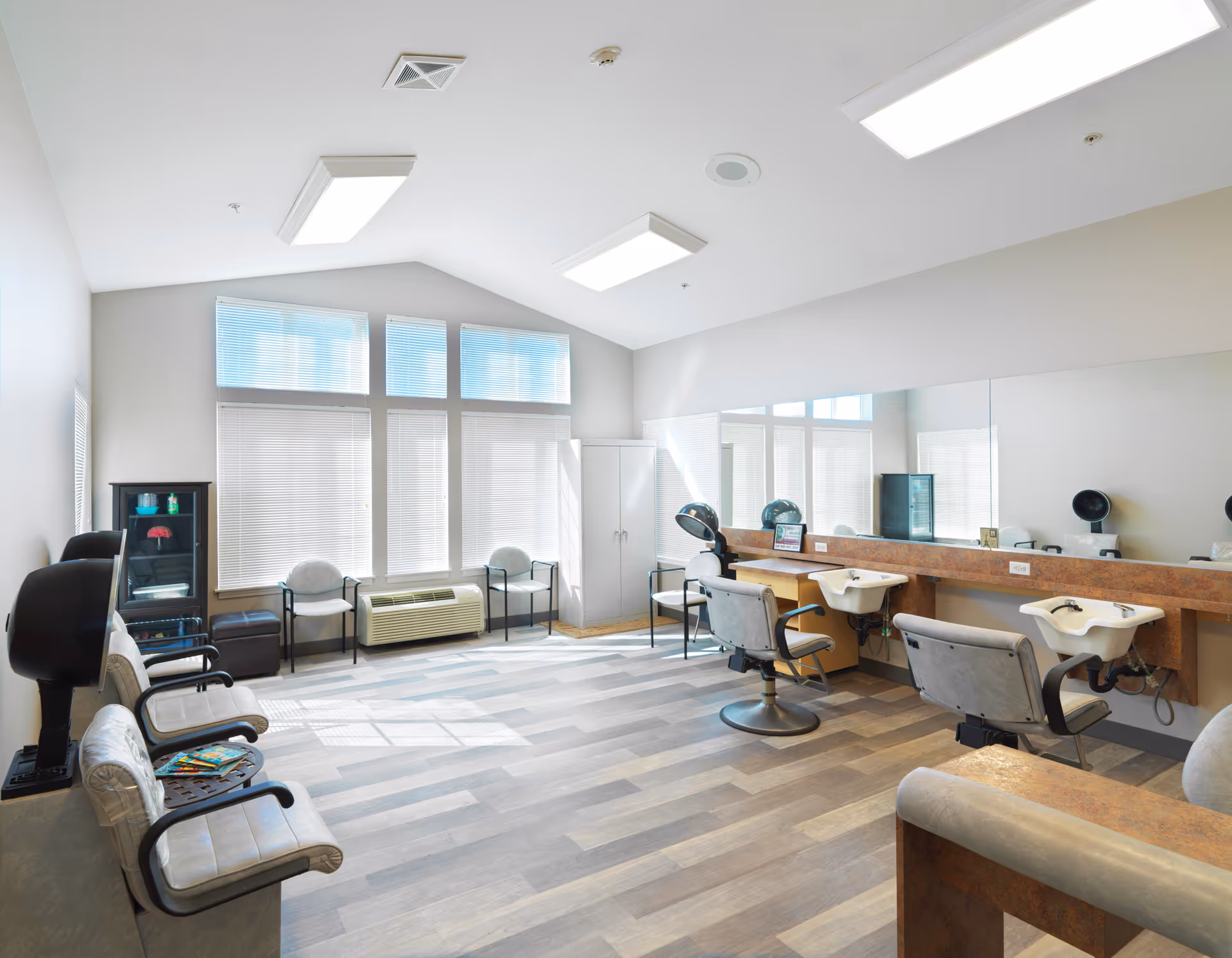 Bright and spacious hair salon area with large windows covered by blinds, multiple salon chairs facing mirrors with hair dryers, and several waiting chairs along the walls. The room has light-colored walls and wood-patterned flooring.