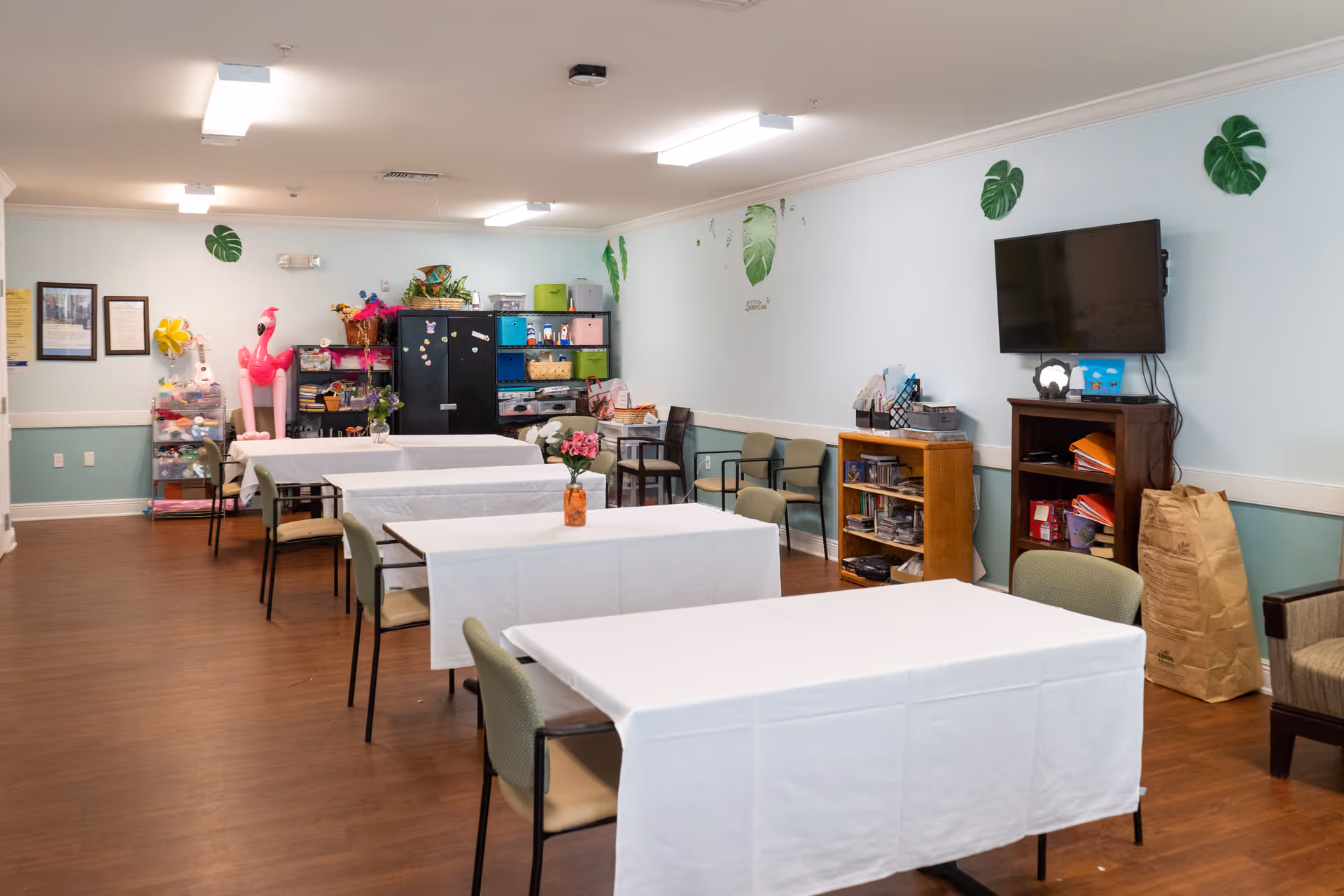 A community room with several tables covered in white tablecloths, each with a small flower arrangement. There are chairs around the tables and along the walls. The walls are light blue with green leaf decorations. A flat-screen TV is mounted on the wall above a wooden shelf filled with books and other items. In the back corner, there are shelves and storage bins, along with a large pink inflatable flamingo. The floor is wood, and the room is well-lit with ceiling lights.