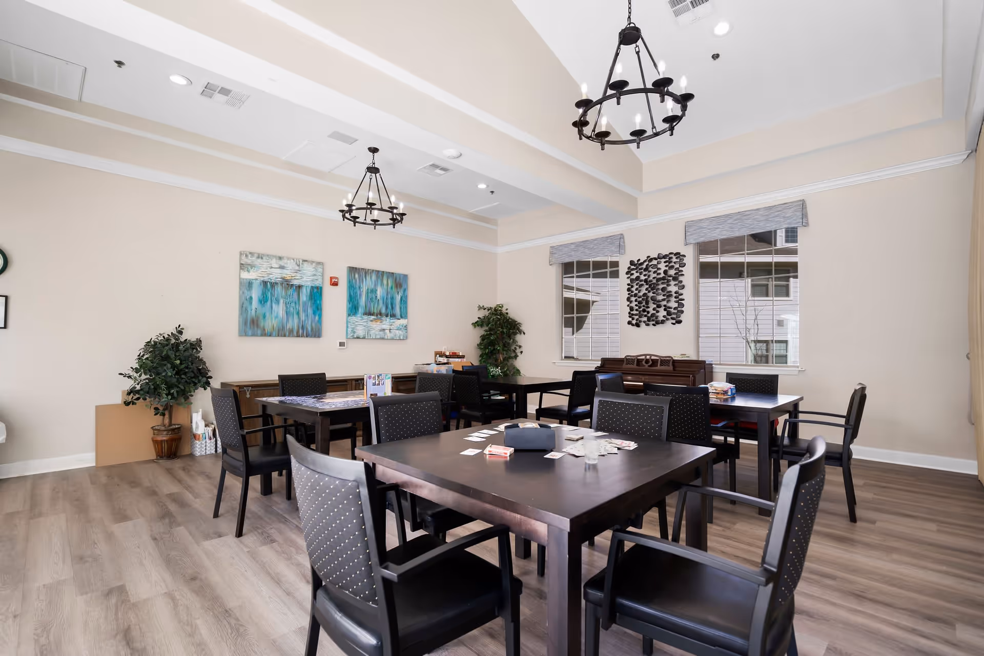 A bright and spacious common room with several dark wooden tables and black chairs arranged for group activities. The room features light-colored walls, two large windows with blinds, decorative wall art, two hanging chandeliers, and a piano against the far wall. The floor is covered with light wood laminate.